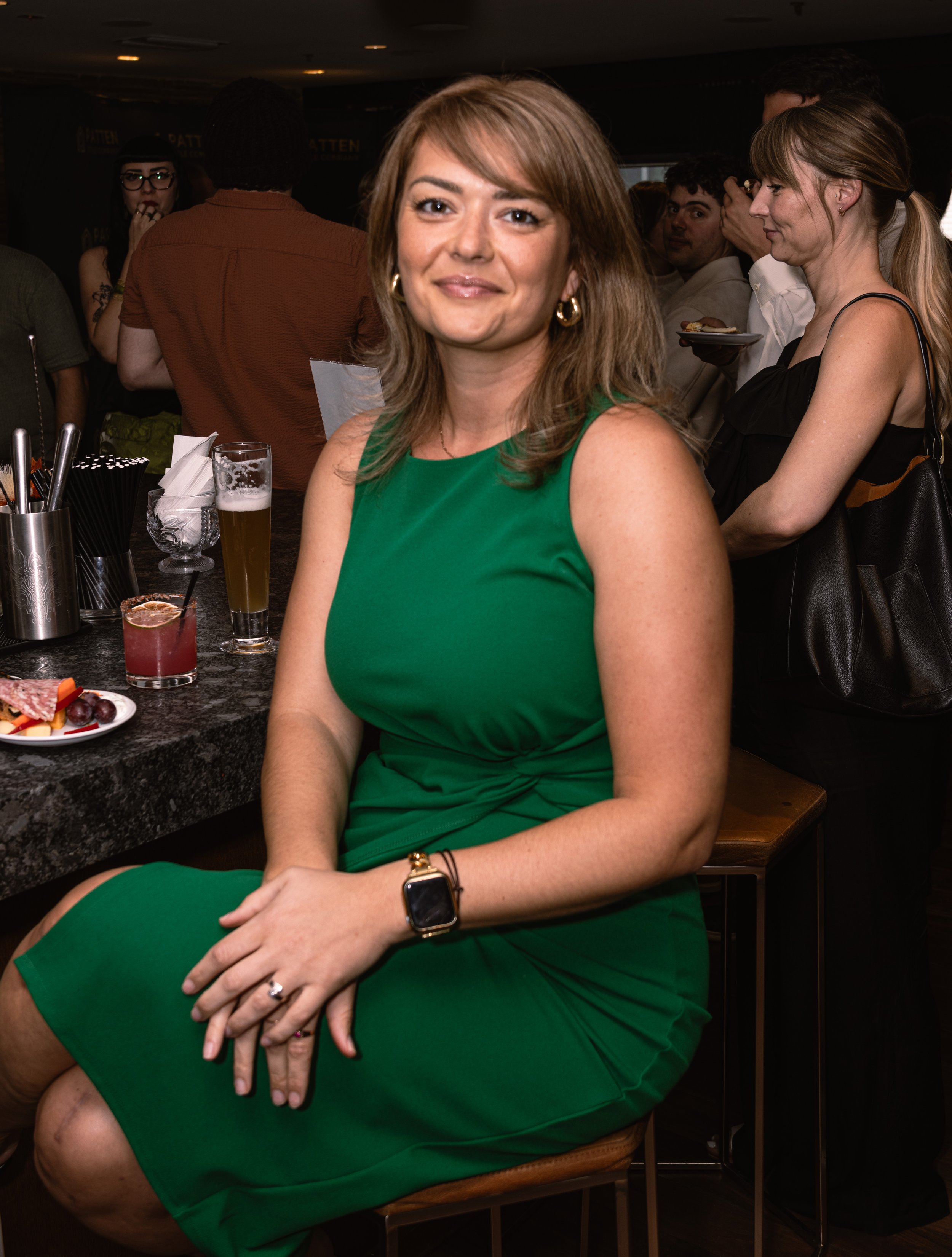A woman in a green dress sitting at a bar, with drinks and snacks in front of her, smiling at the camera, in a crowded indoor setting.