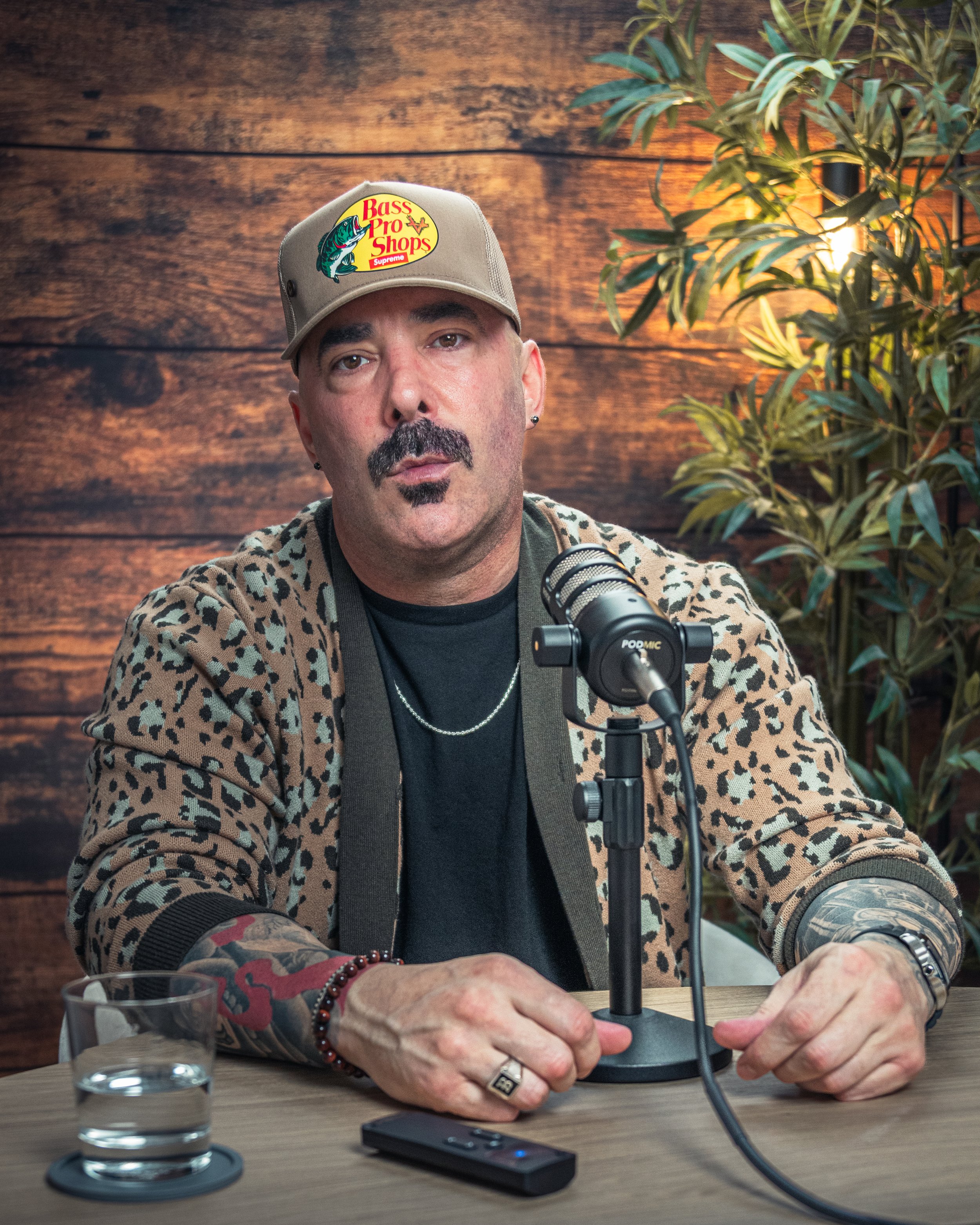 A person wearing a tan cap with a Bass Pro Shops logo, a leopard print jacket, and various jewelry, seated at a table with a microphone in front of them. Their right hand is near a small device on the table. In the background, there is a wooden wall 