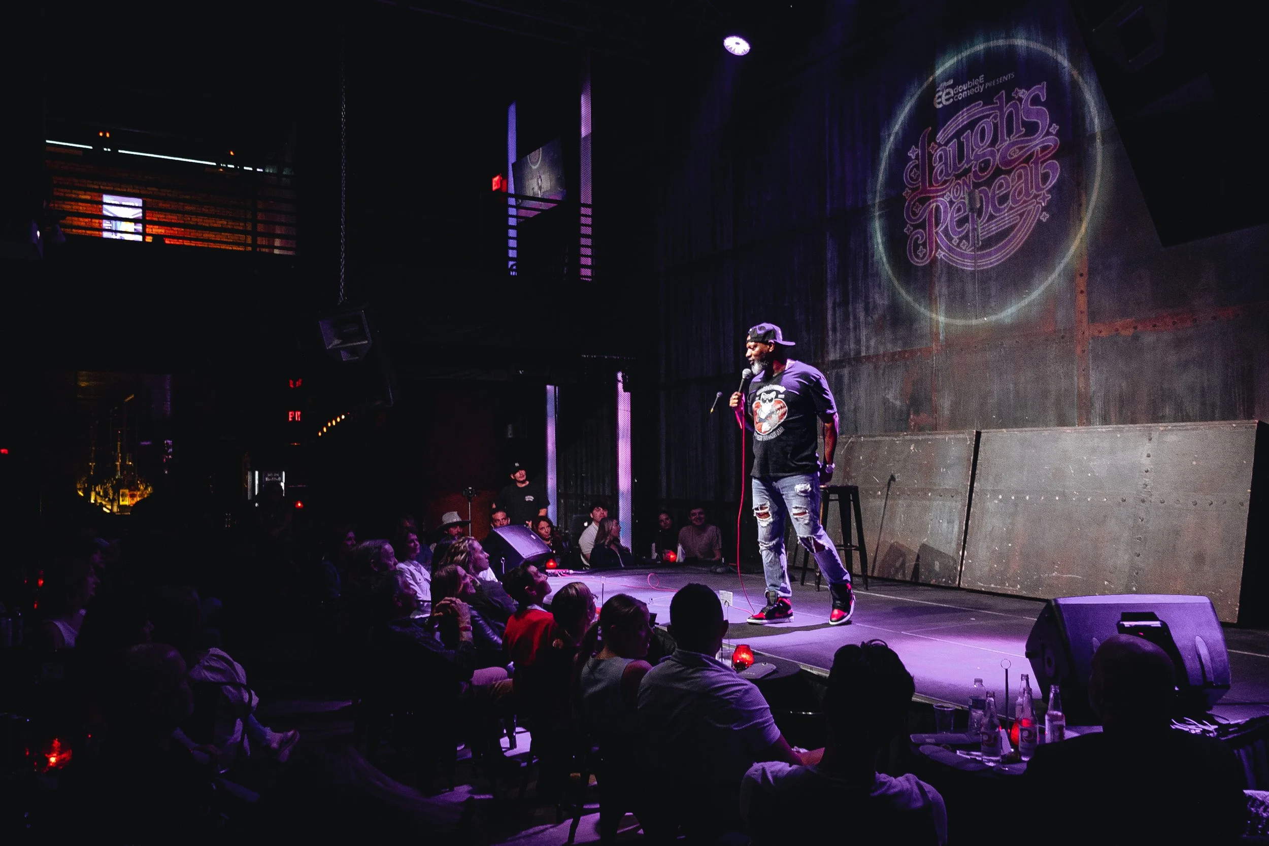 A stand-up comedian performs on stage at a comedy show titled 'Laughs on Rebar' in a dimly lit venue with an audience seated and watching. The comedian is wearing a hat, glasses, and a graphic T-shirt, holding a microphone.