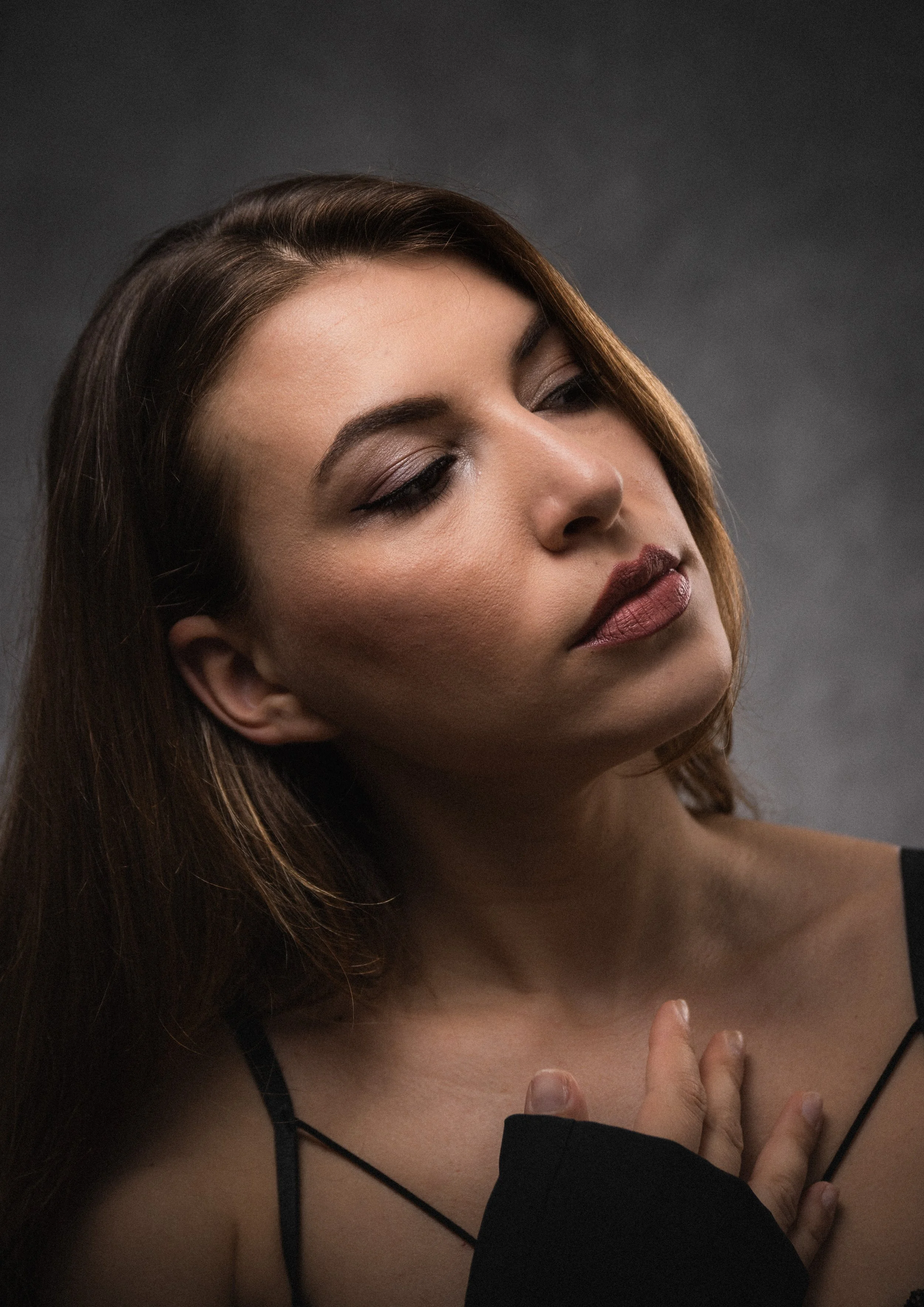 A young woman with long brown hair and makeup wearing a black strapless top and black sheer sleeve, with her hand resting on her chest against a dark gray background.