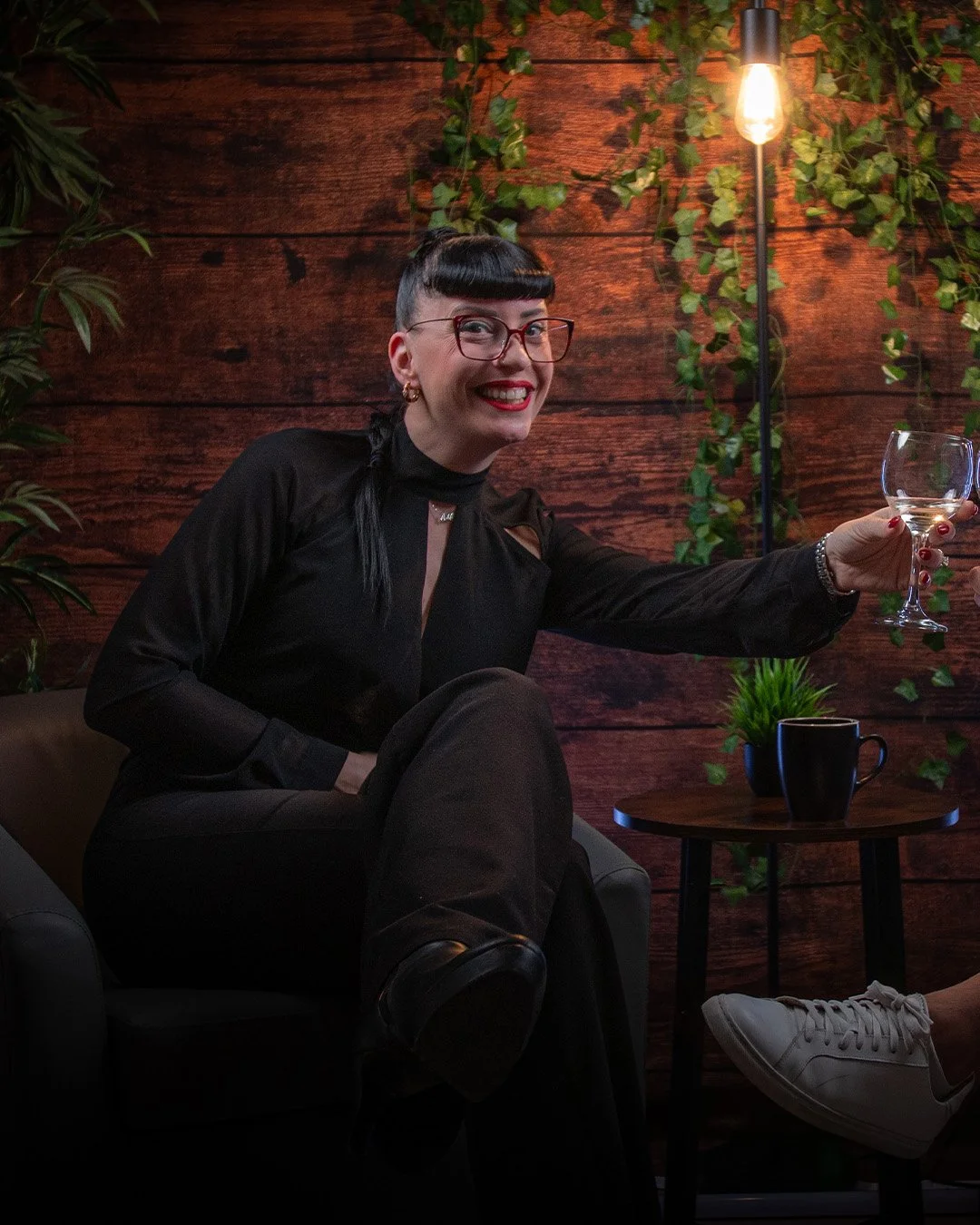 A smiling woman with black hair and glasses sitting in a cozy room with wooden walls and green plants, holding a wine glass in her extended hand, with a small table and a plant in a pot beside her.