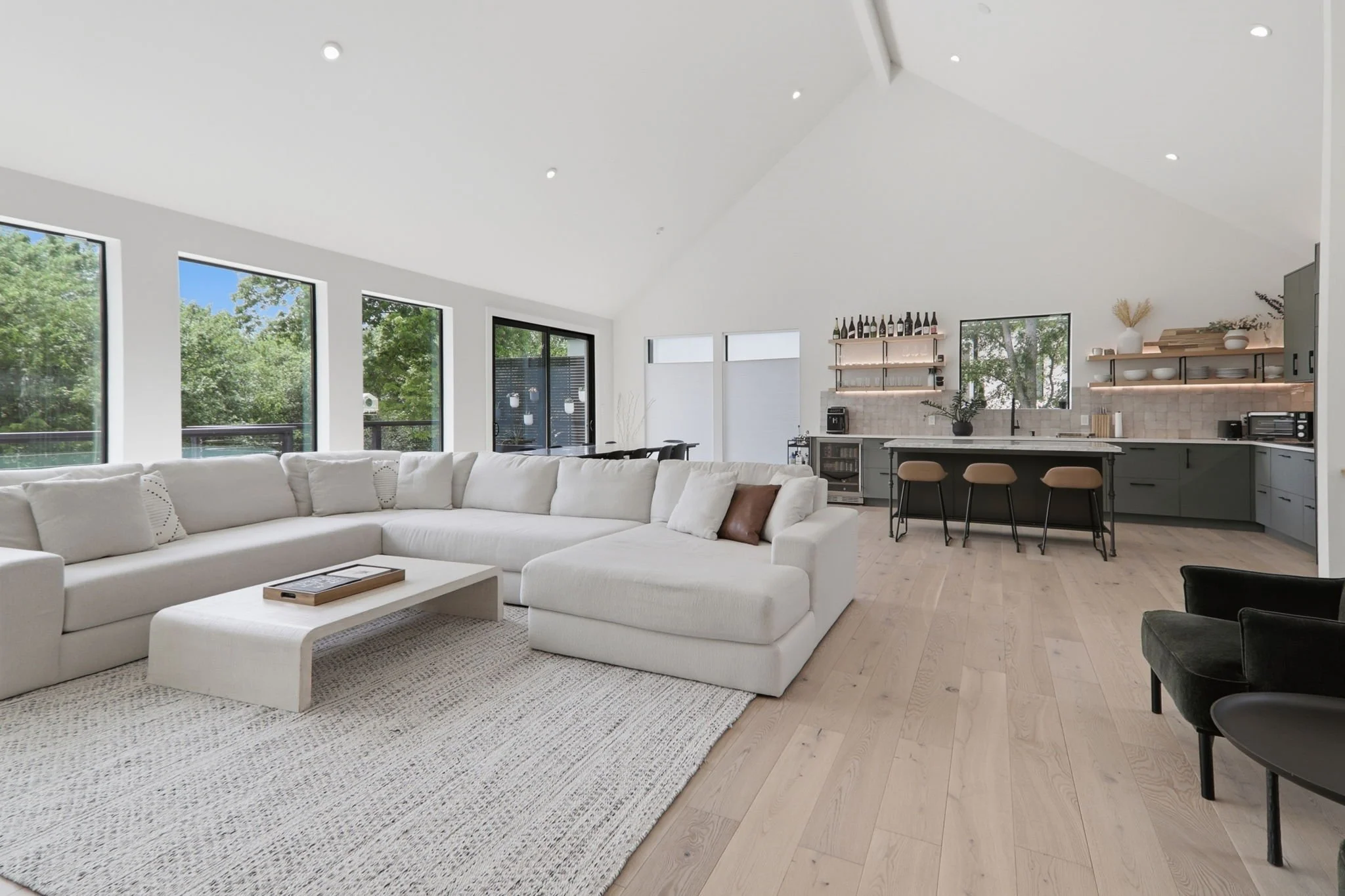 A modern open-concept living room and kitchen with large windows, white walls, light wood flooring, a white sectional sofa, a coffee table, and a kitchen area with open shelves, bar stools, and various decor items.