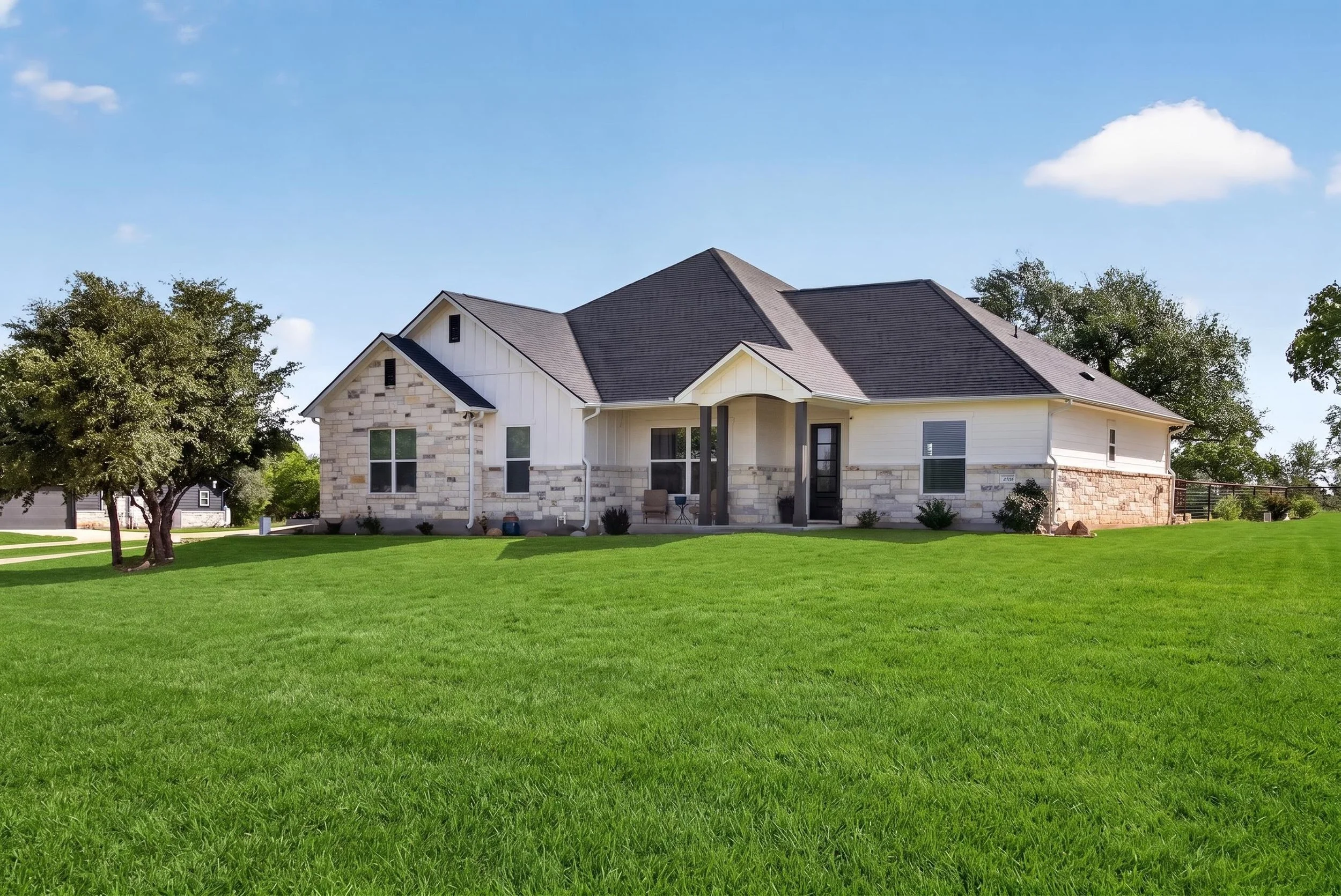 A modern suburban house with a well-maintained green lawn and a large tree in the front yard, under a blue sky with some clouds.