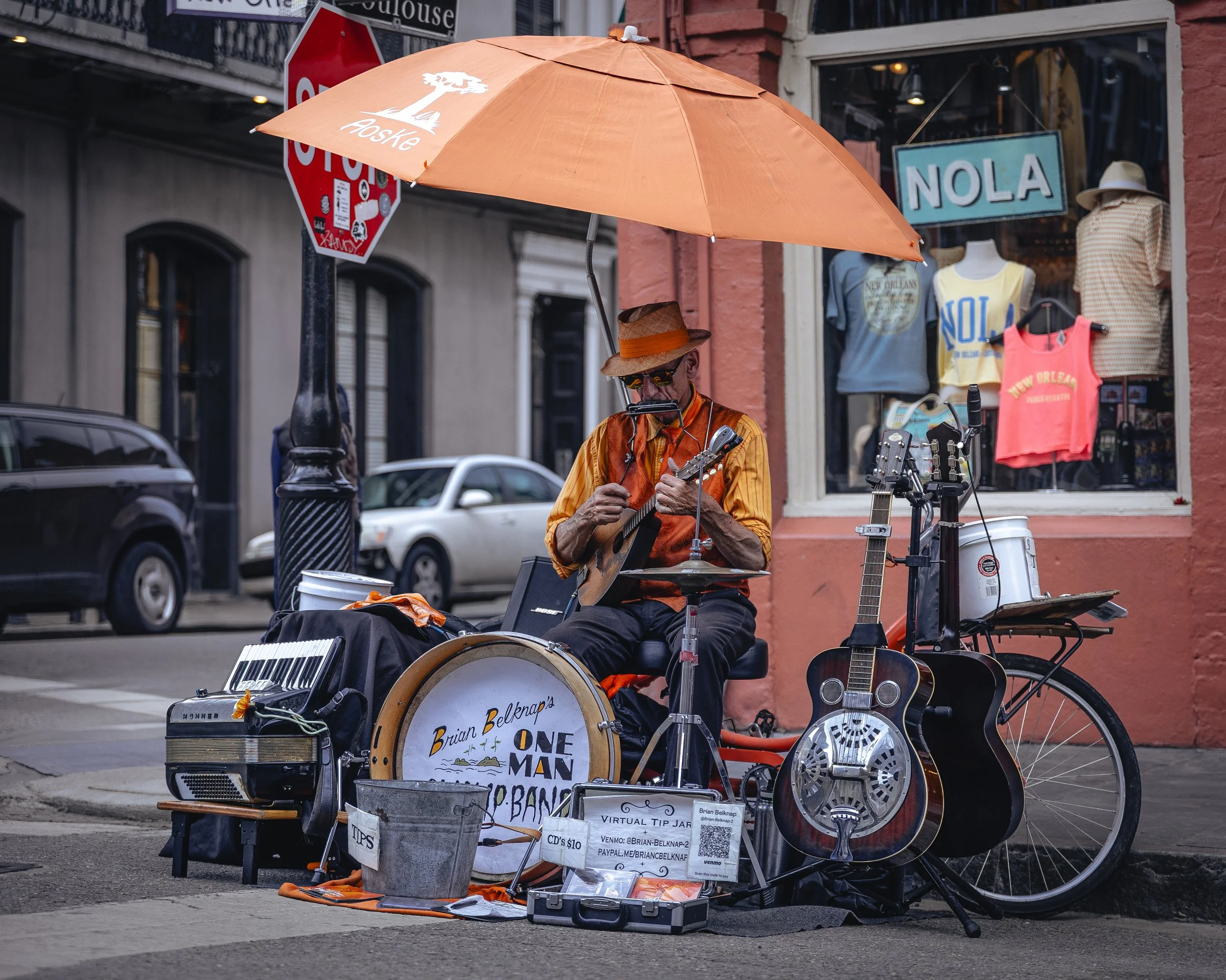 A street performer sitting under an orange umbrella with a sign reading 'Brian Belknap One Man Band,' surrounded by musical equipment and donation containers, on a city sidewalk in front of a clothing store with mannequins wearing T-shirts and tank t