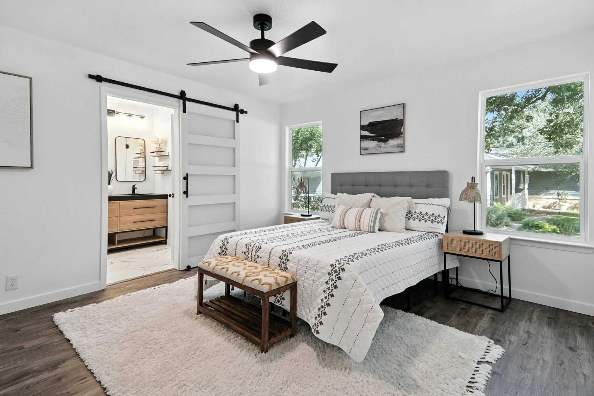 A bedroom with white walls, large windows showing green trees outside, a bed with black and white patterned bedding, two bedside tables with lamps, a framed black and white landscape photo on the wall, a cream-colored bench at the foot of the bed, a 