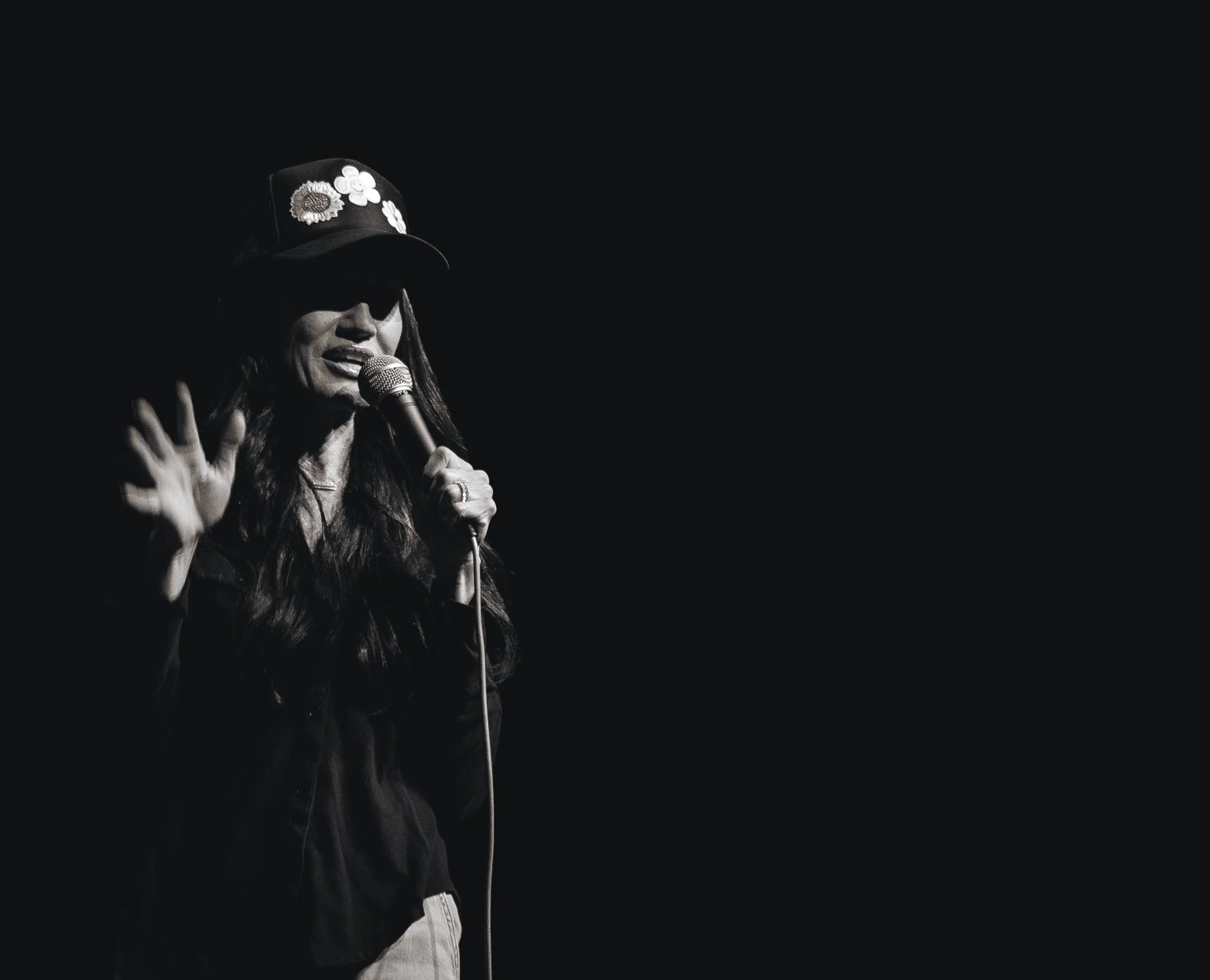 Black and white photo of a woman wearing sunglasses and a cap with flowers, singing into a microphone.