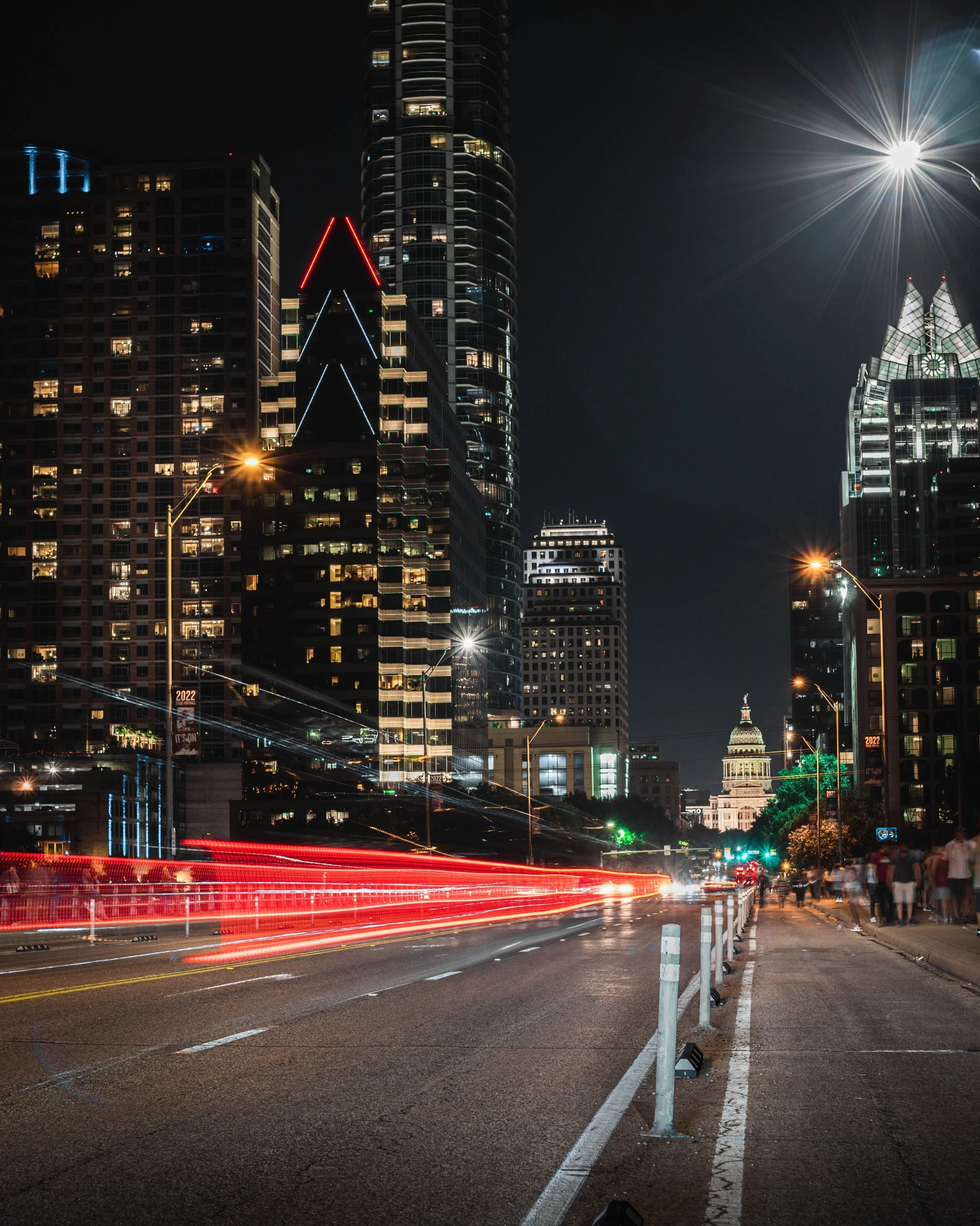 Nighttime cityscape with tall illuminated skyscrapers, moving vehicle light trails, and the United States Capitol building in the distance.