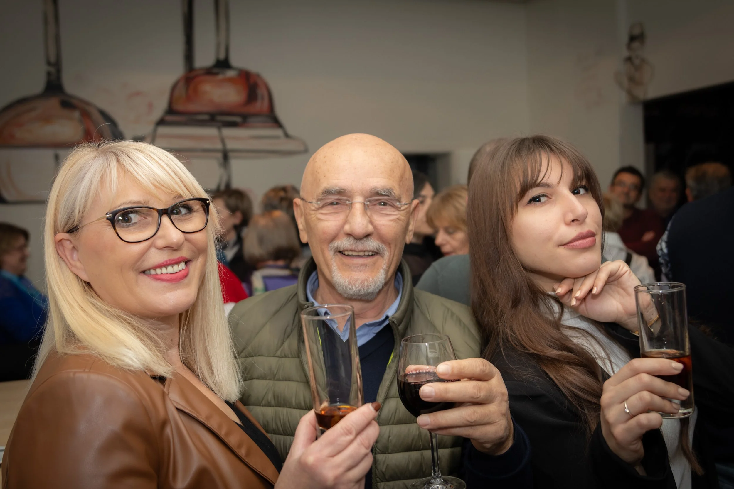 Three people smiling and holding glasses of wine at a social event, with a busy background of other guests and wall art.