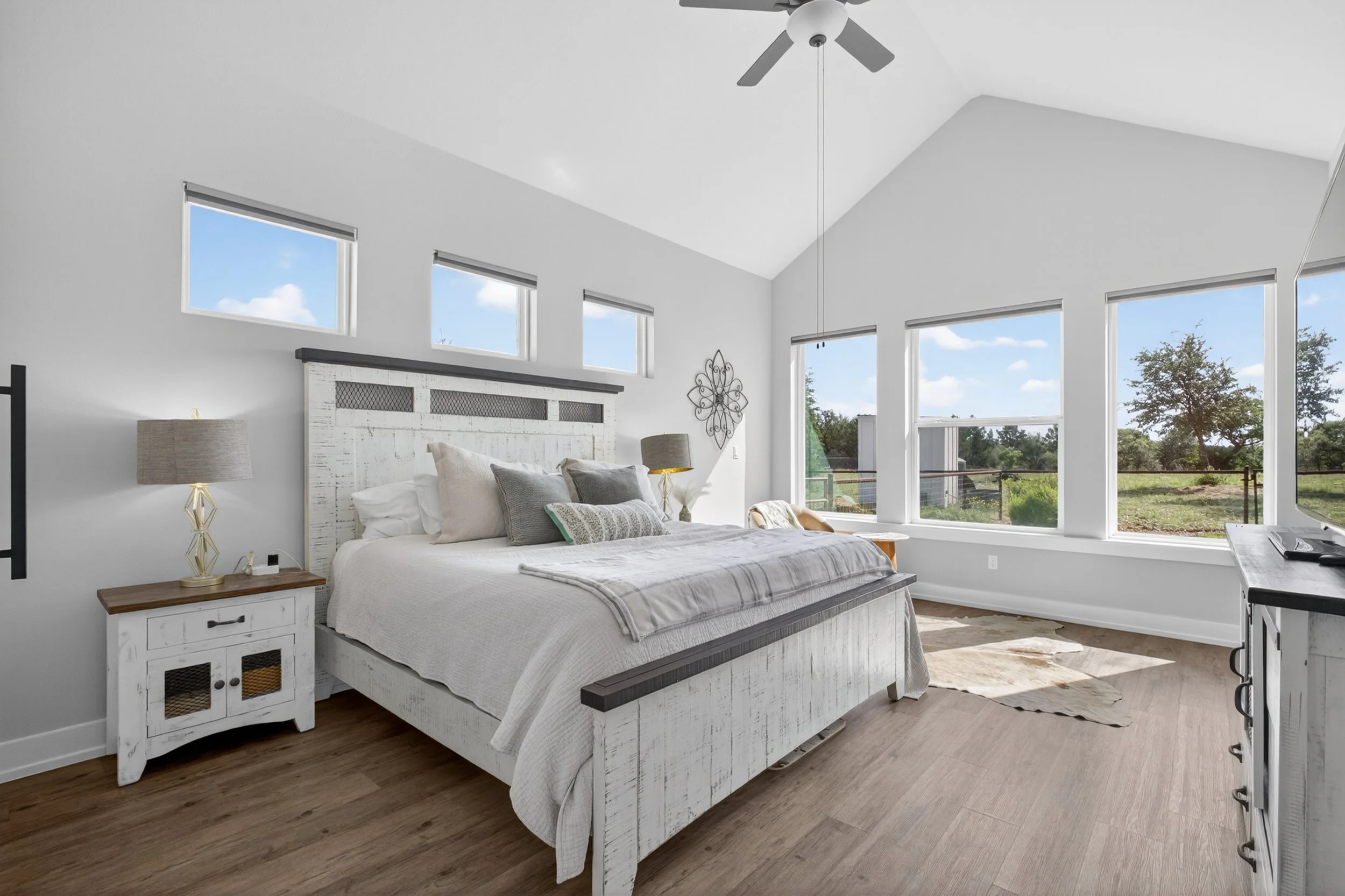 Bright bedroom with high ceilings, large windows showing a blue sky and green trees, a white distressed bed with multiple pillows, matching white distressed side table with a lamp, and wood flooring.