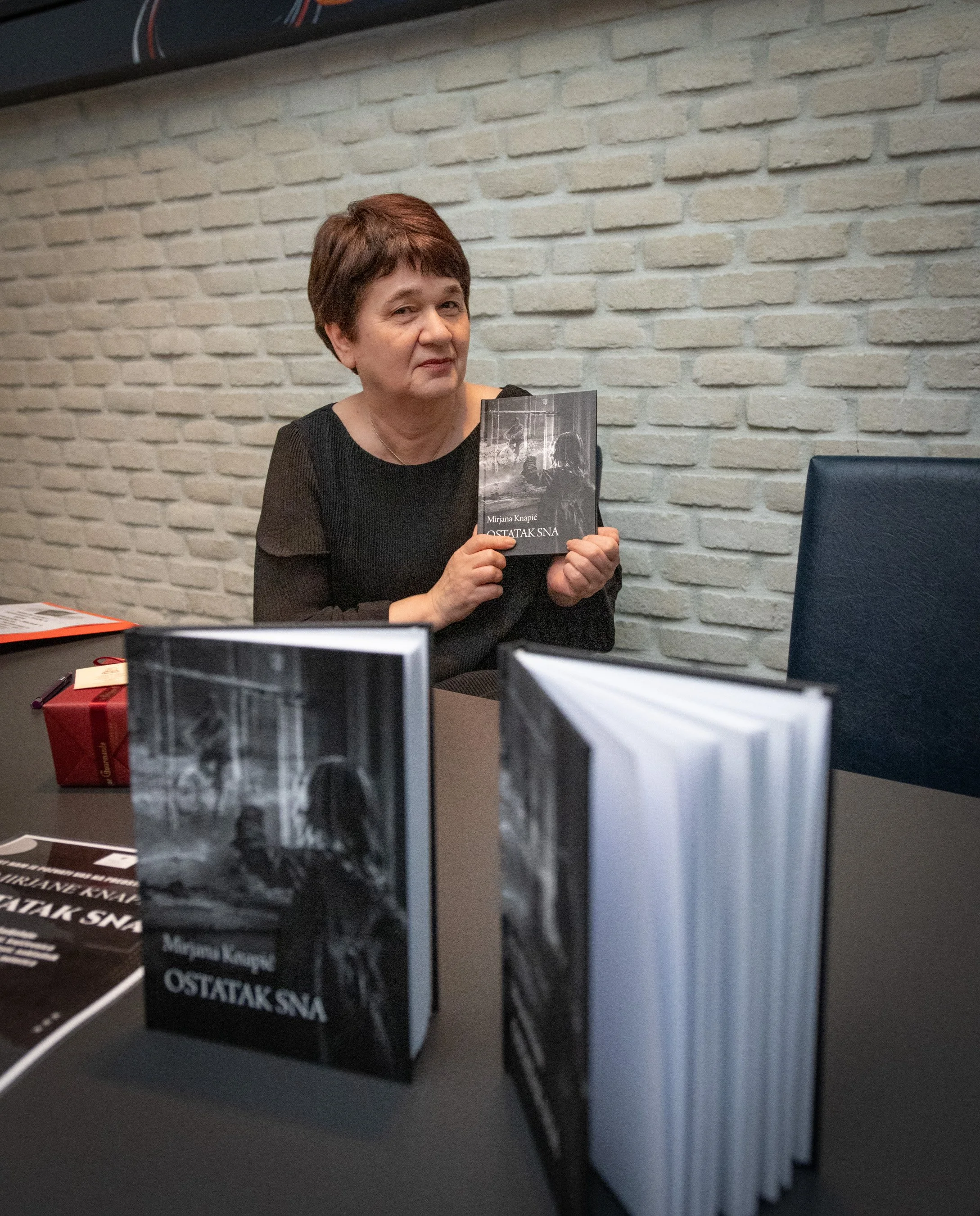 A woman with short brown hair sitting at a table holding a book titled "OstavaK SNA" by Mirjana Knapic. The background has a white brick wall, and there are books and a red gift box on the table.