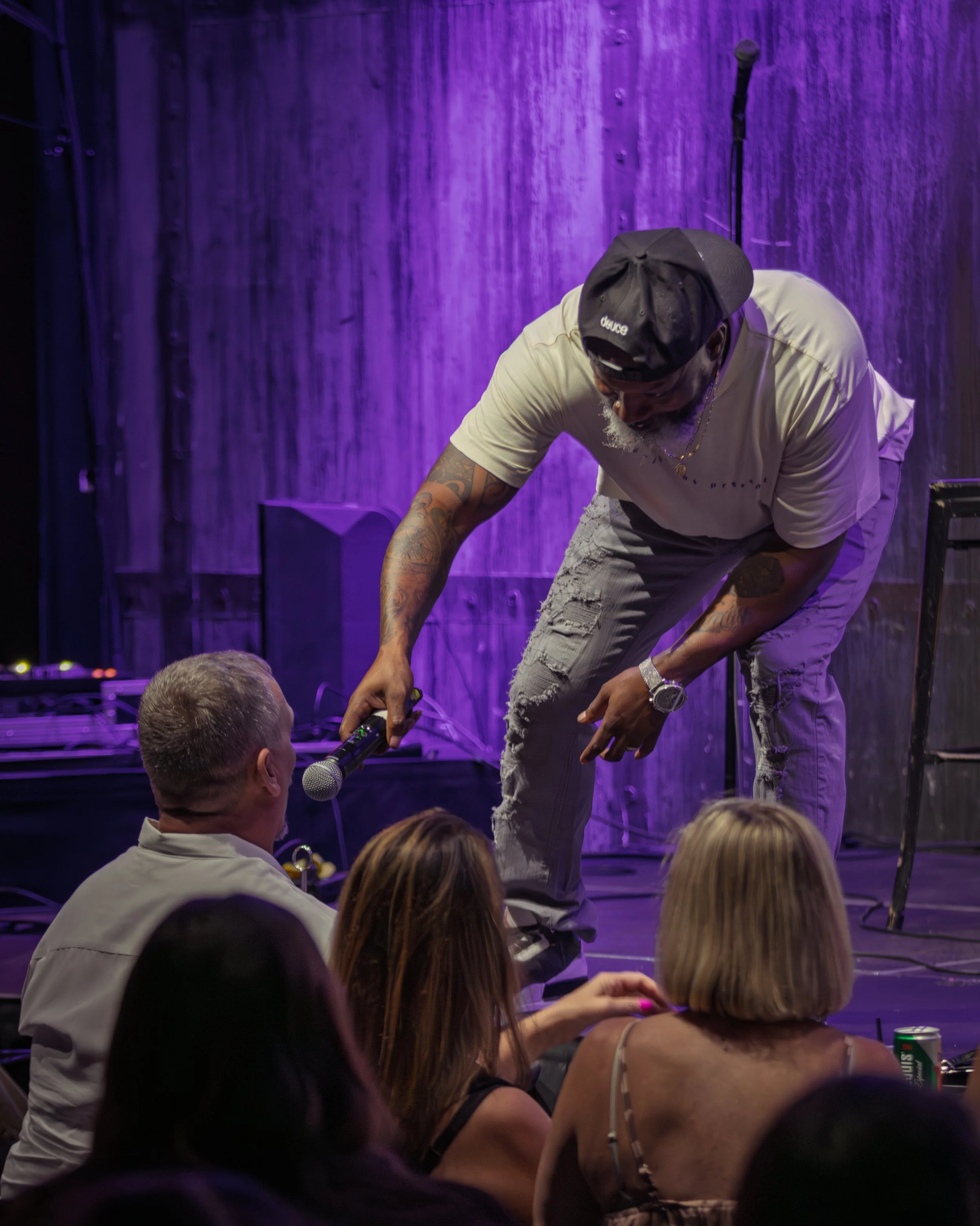 A man in a beige t-shirt, ripped jeans, black cap, and jewelry bends down to speak into a microphone held by an audience member at a performance or event. The background features a purple-lit wooden wall and a standing microphone.