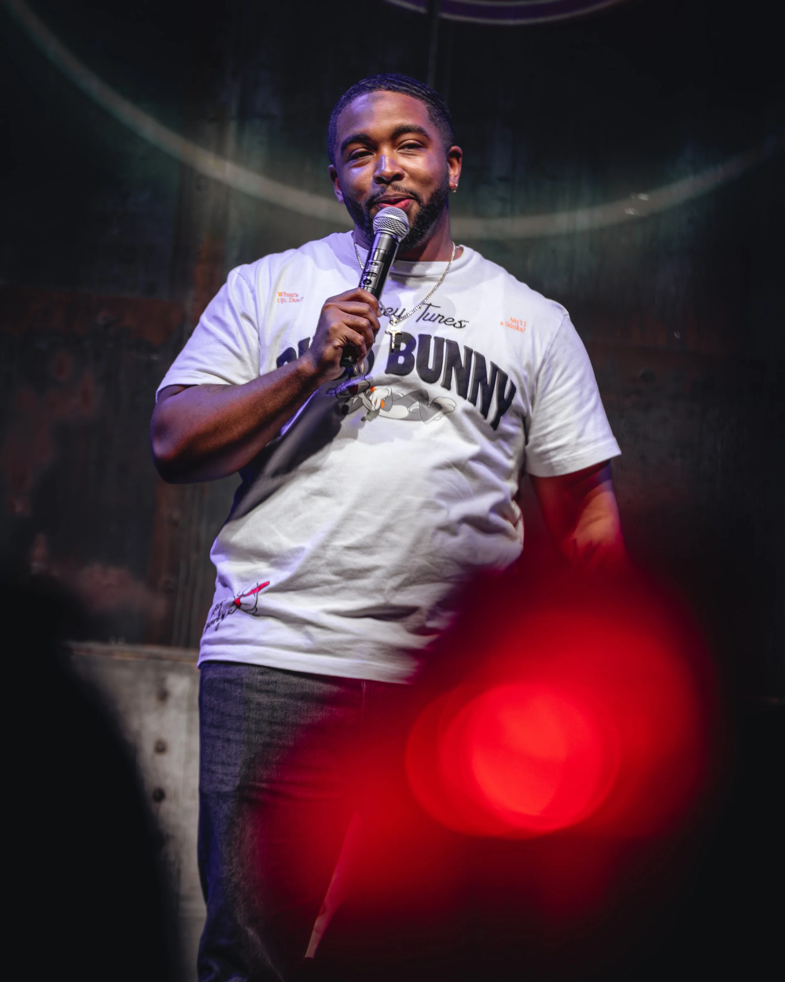 A man with short hair and a beard is holding a microphone on stage, smiling. He's wearing a graphic T-shirt with the word 'BUNNY' and cartoon bunny images, and a silver chain necklace. The background is dark with red lighting effects.