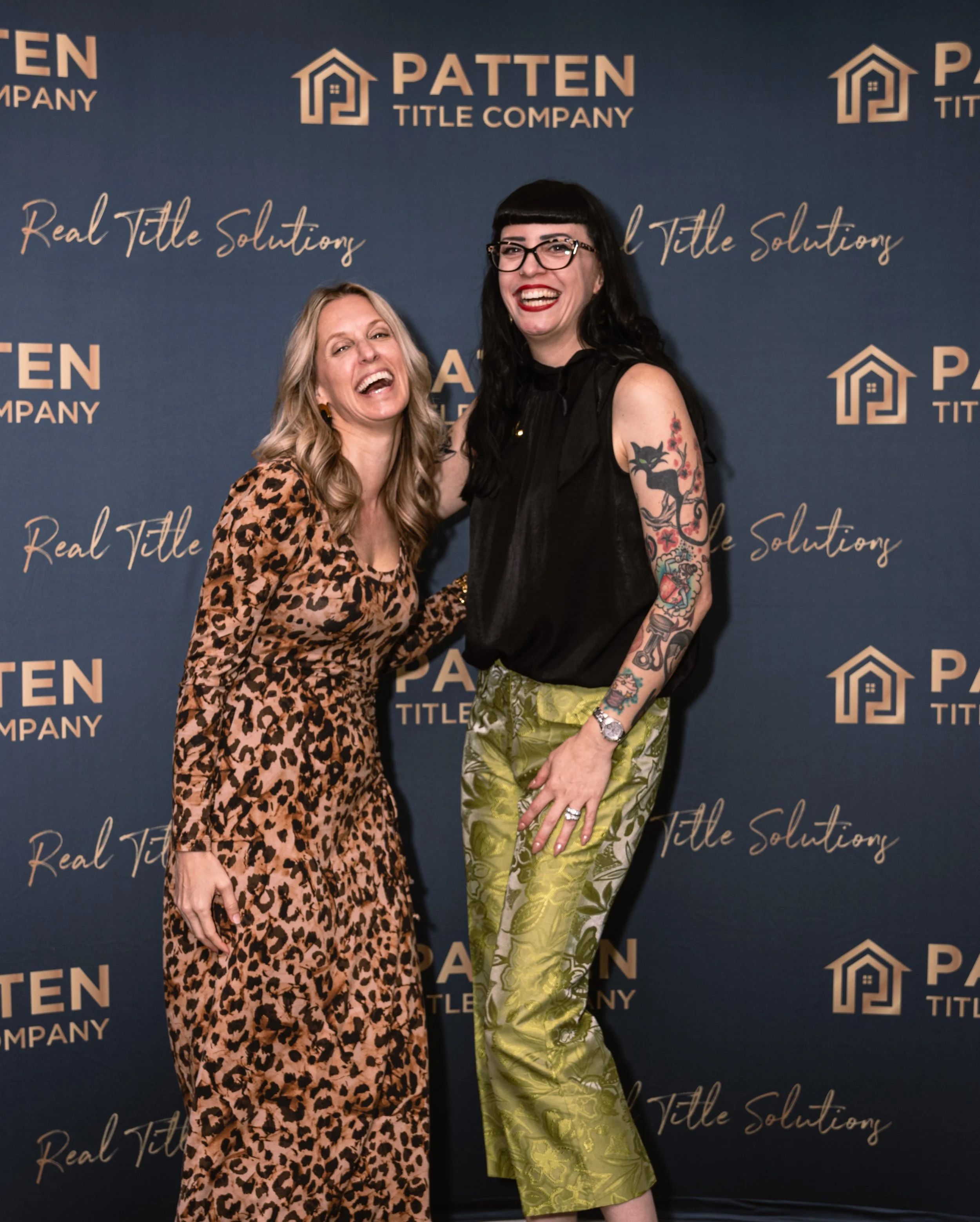Two women smiling and posing in front of a step and repeat banner with the logos and text of Patten Title Company. The woman on the left has blonde hair, wearing a leopard print dress. The woman on the right has black hair, glasses, tattoos on her ar