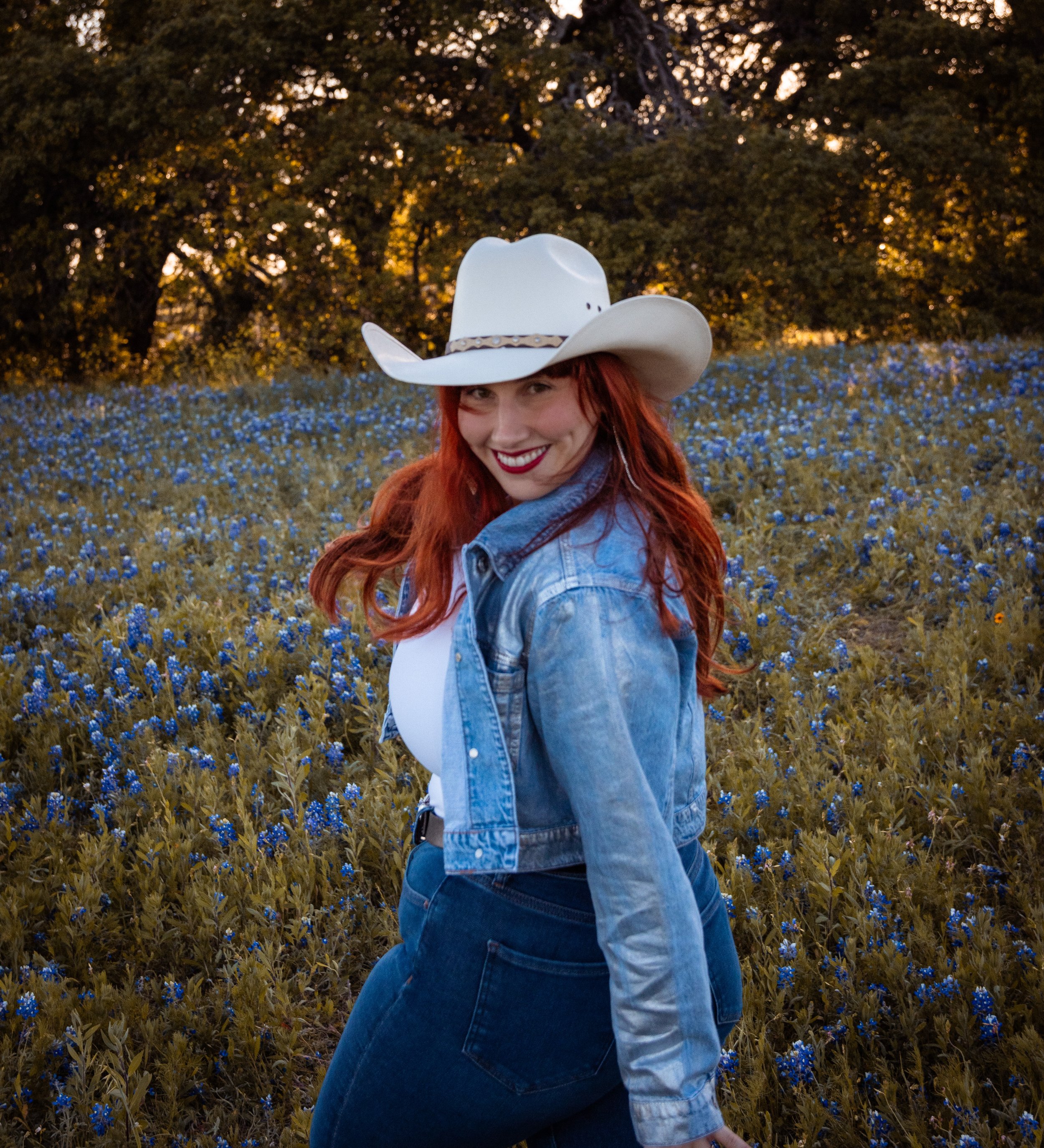 A young woman with red hair, wearing a large white cowboy hat, a denim jacket, and jeans, standing in a field of blue flowers with trees in the background and sunlight shining through.