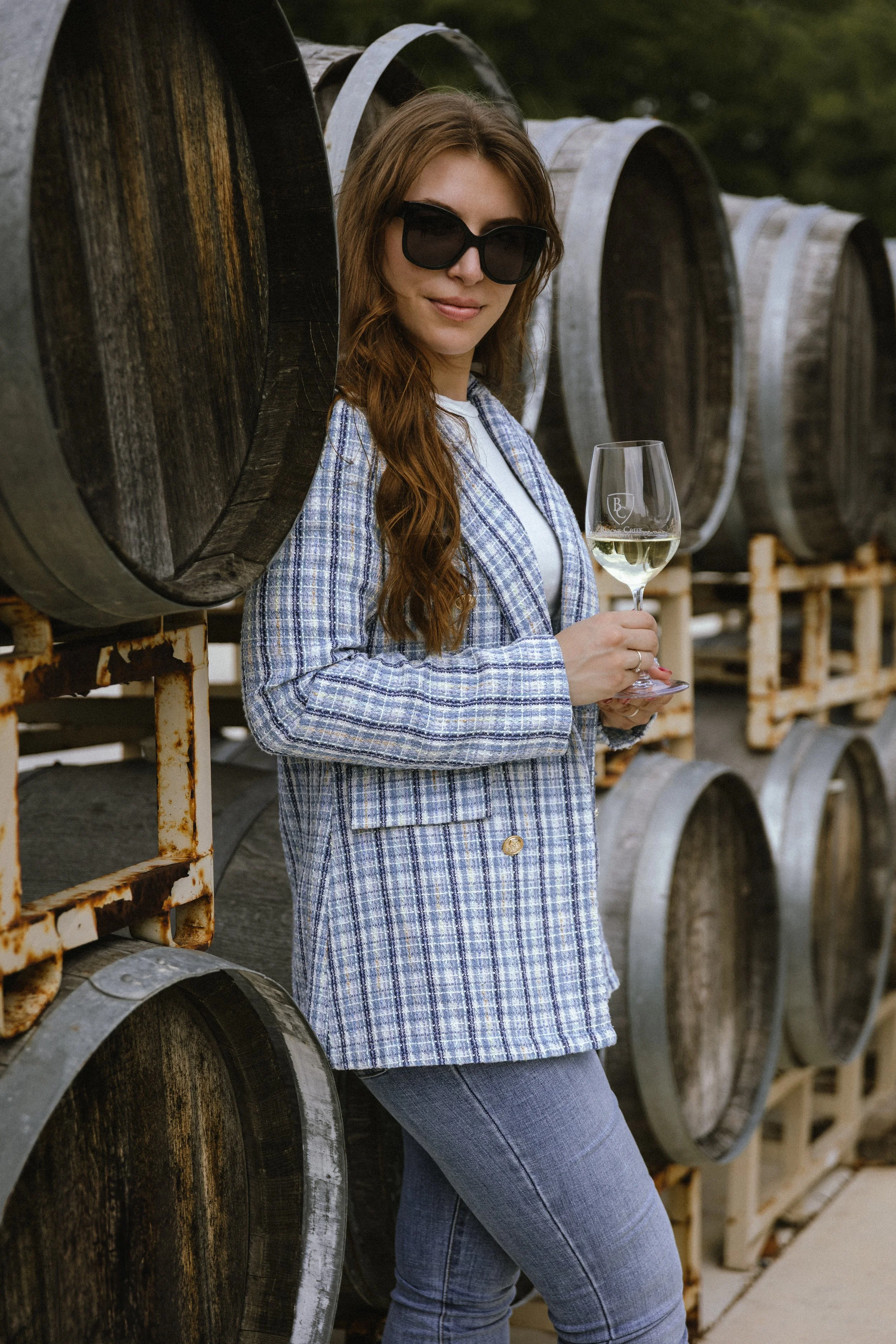 A woman wearing sunglasses and a light blue plaid jacket holding a glass of white wine, standing beside large oak barrels in a winery or wine cellar.