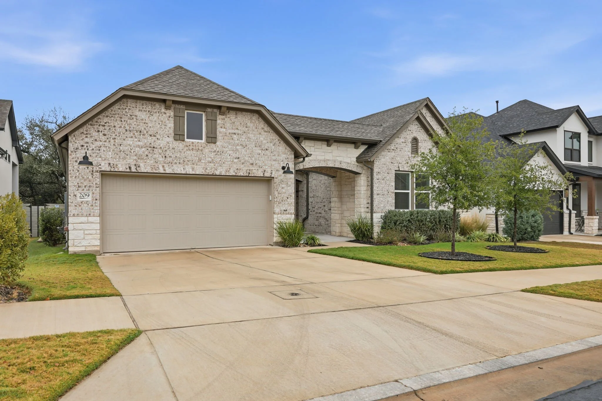 The front view of a modern house with brick and stucco exterior, a two-car garage, paved driveway, small trees, and landscaped yard, under a blue sky.