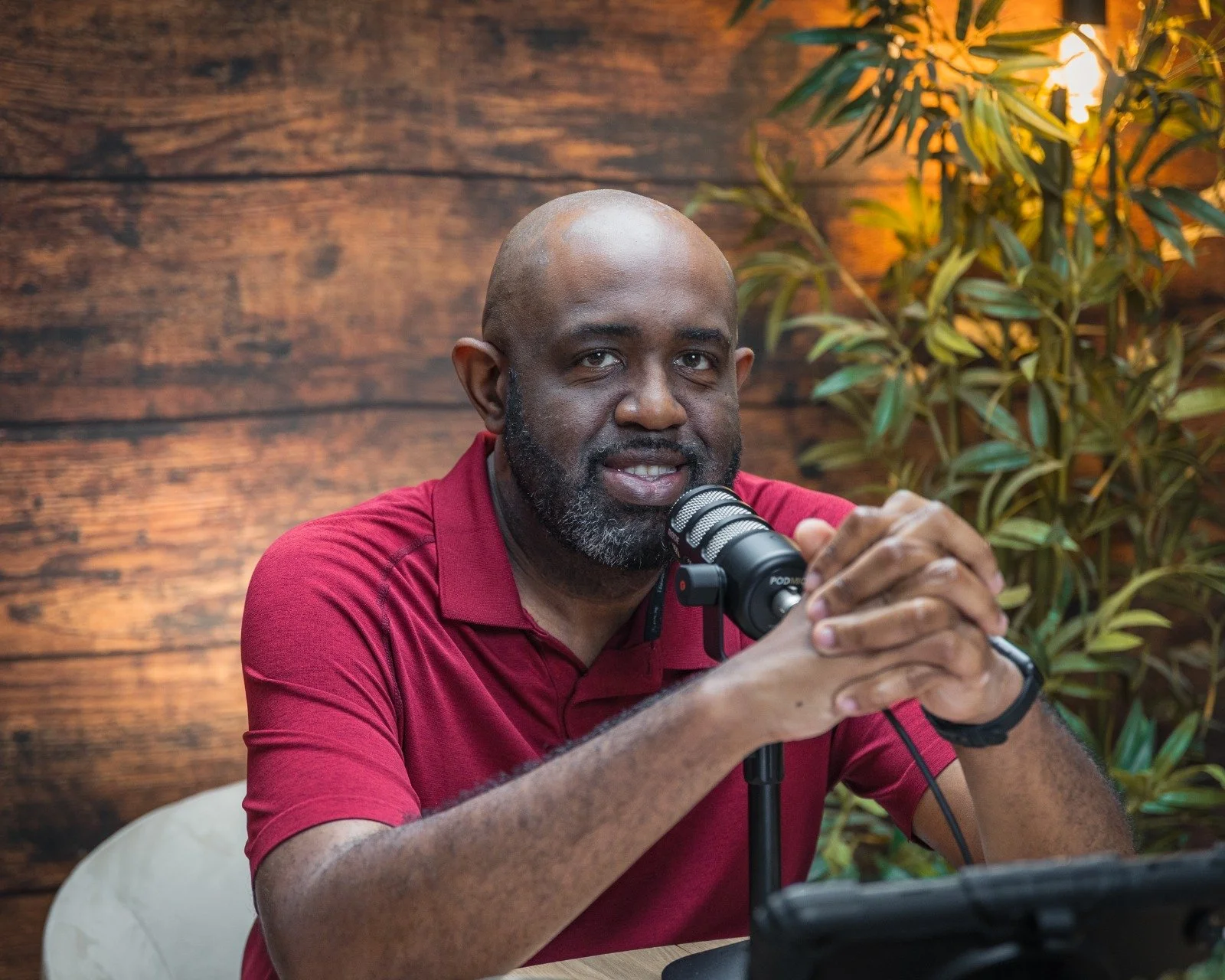 A man with a bald head and beard, wearing a red polo shirt, sitting at a table with a microphone in front of him, speaking or smiling. Background features wooden wall panels and a potted plant.