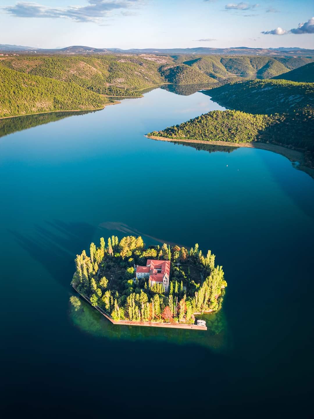 An aerial view of a large body of water with a small, tree-covered island featuring a house or building with red roofs and surrounding lush greenery. Hills and mountains are visible in the background under a partly cloudy sky.