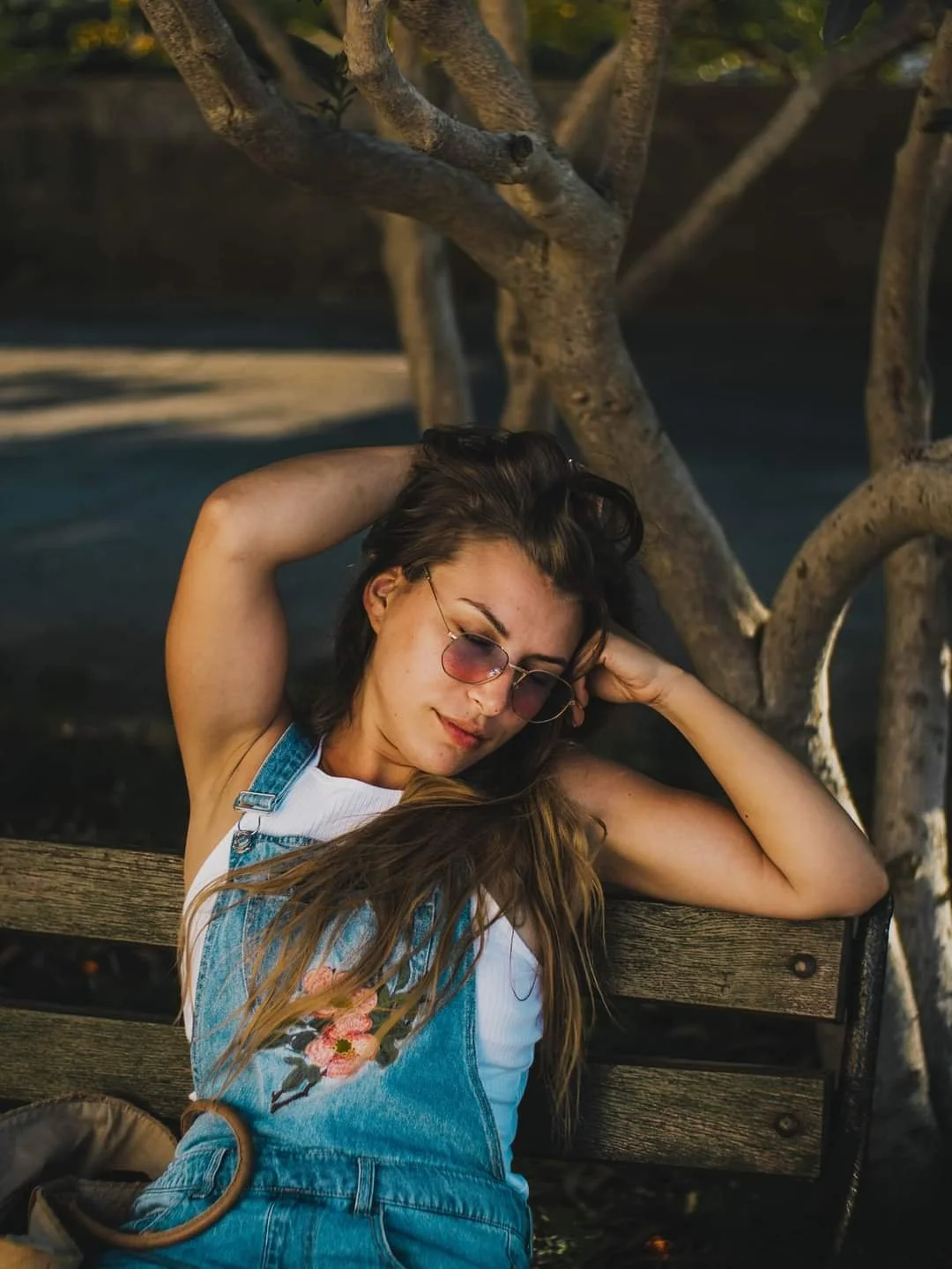 Young woman with sunglasses and long hair lying on a park bench with her arms behind her head, relaxing near a tree.