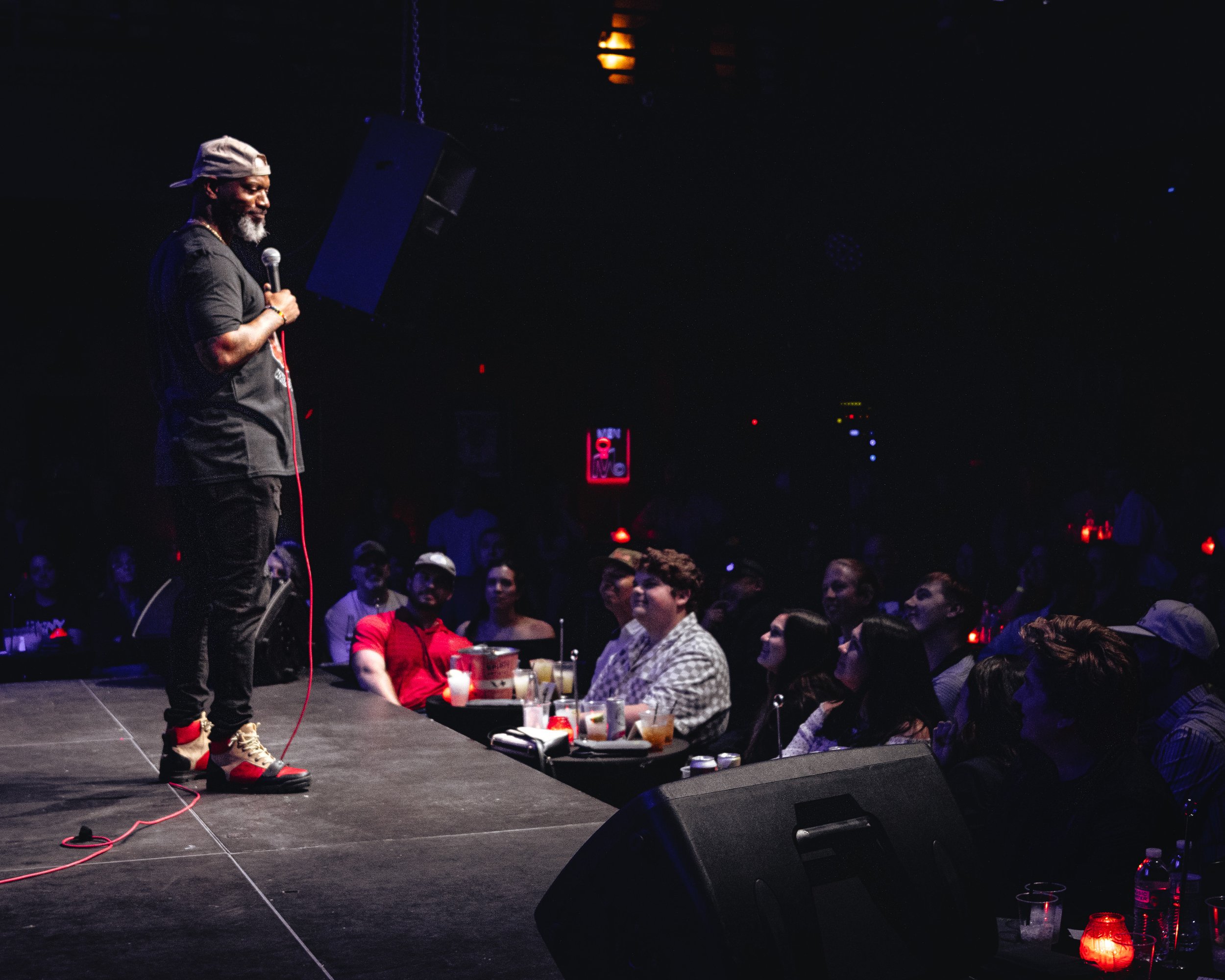 A man in a black shirt, gray cap worn backwards, and beige sneakers holding a microphone on stage during a comedy show, with an audience seated at tables in a dimly lit venue.