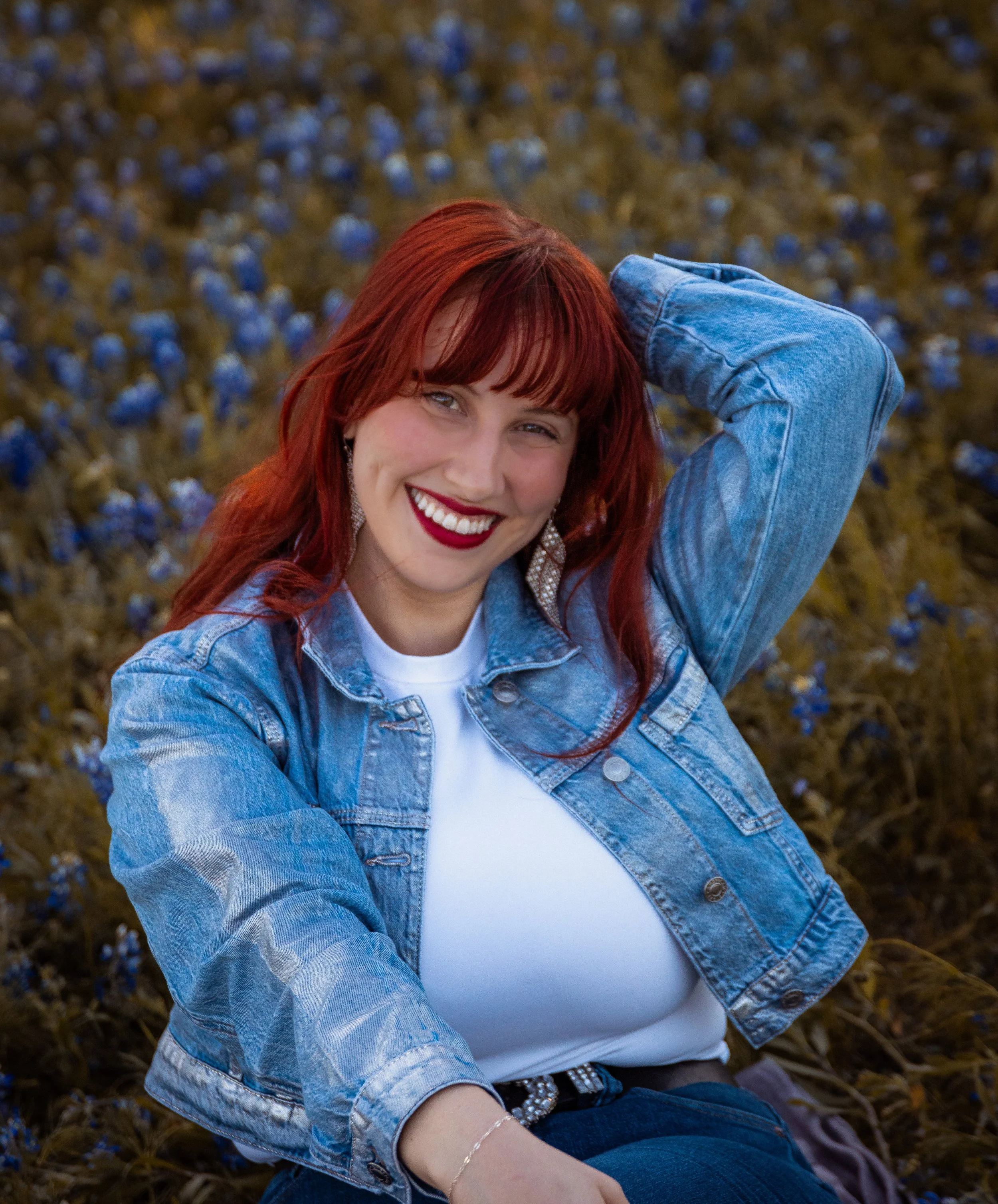 Close-up of a woman with red hair smiling, wearing a denim jacket and white top, sitting outdoors in a field of blue flowers.