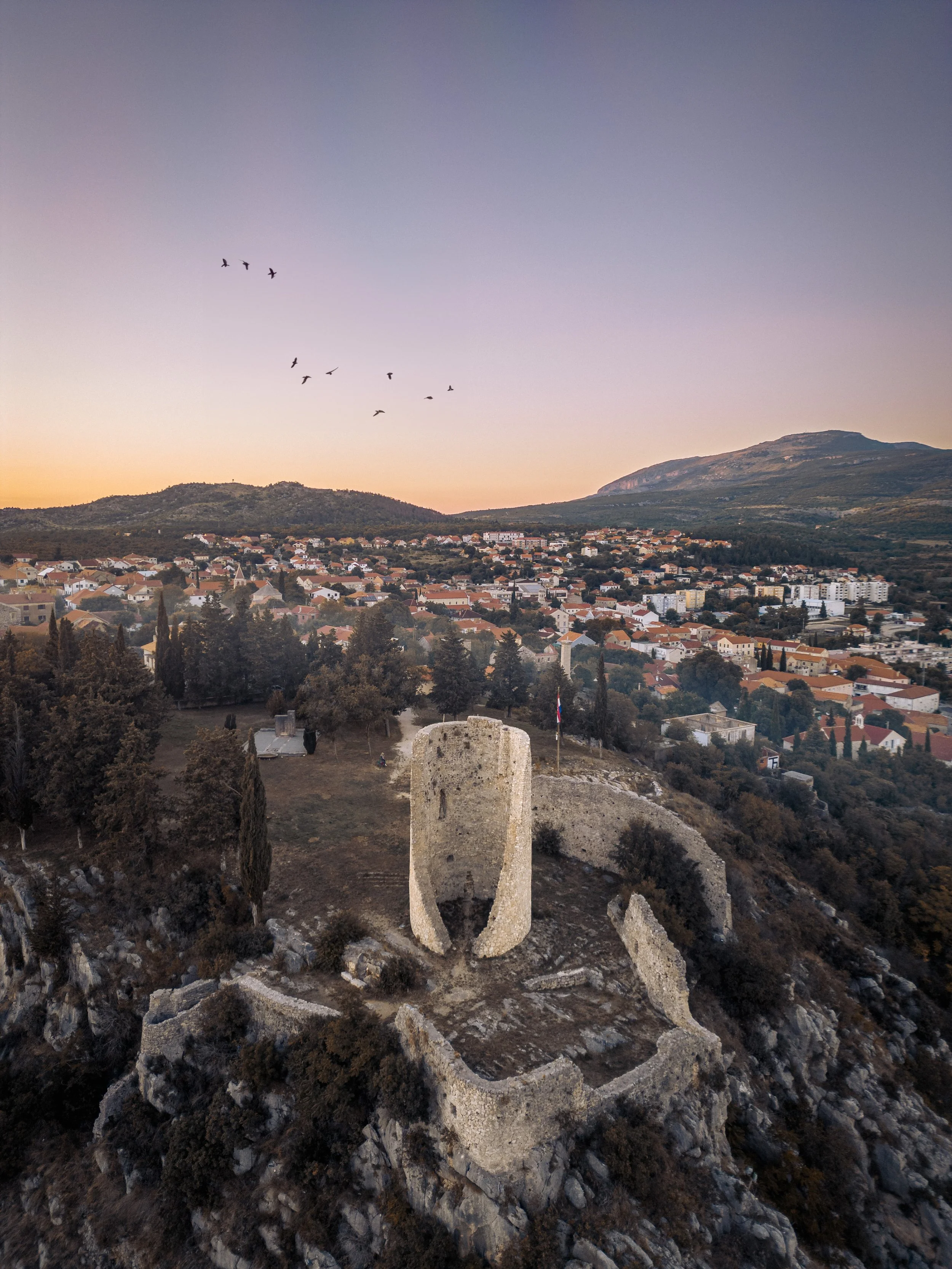 Archaeological ruins on a hill with rocks and stones, overlooking a town with houses and a mountain in the background, during sunset.