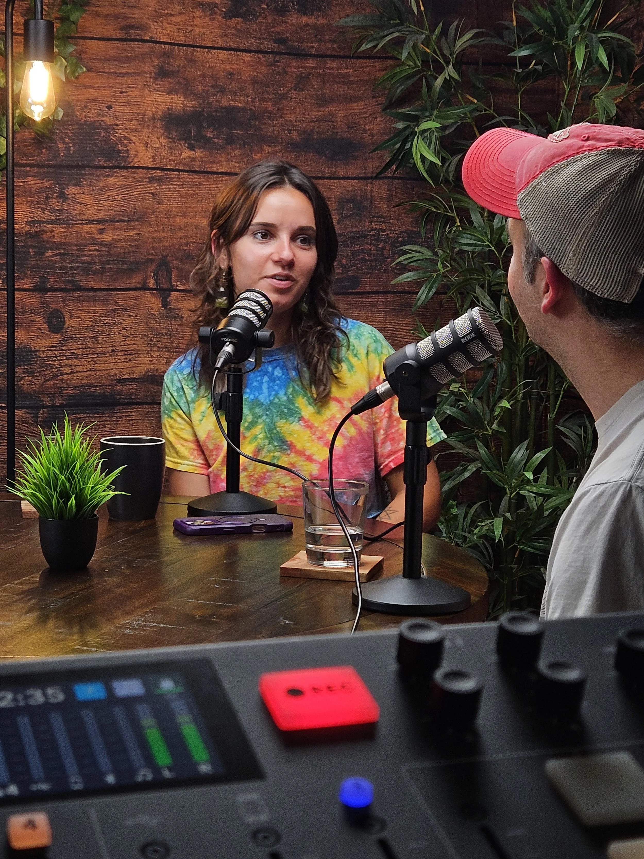 Two people having a conversation with microphones in front of them, seated at a wooden table with a potted plant, a glass of water, and a cell phone, against a background of wooden wall with green plants.