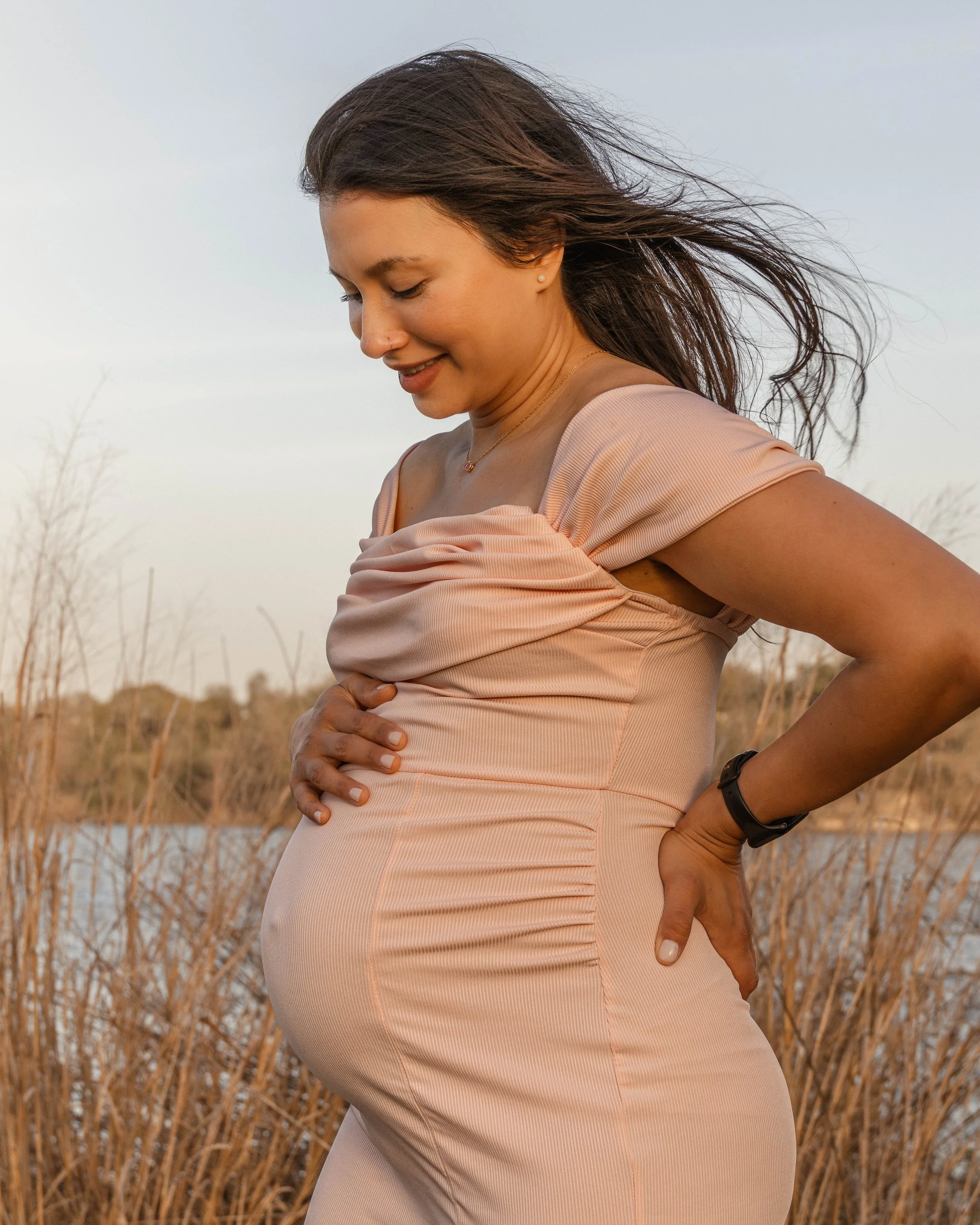 Side view of a pregnant woman with long brown hair blowing in the wind, standing outdoors in a field with dry plants and a cloudy sky.