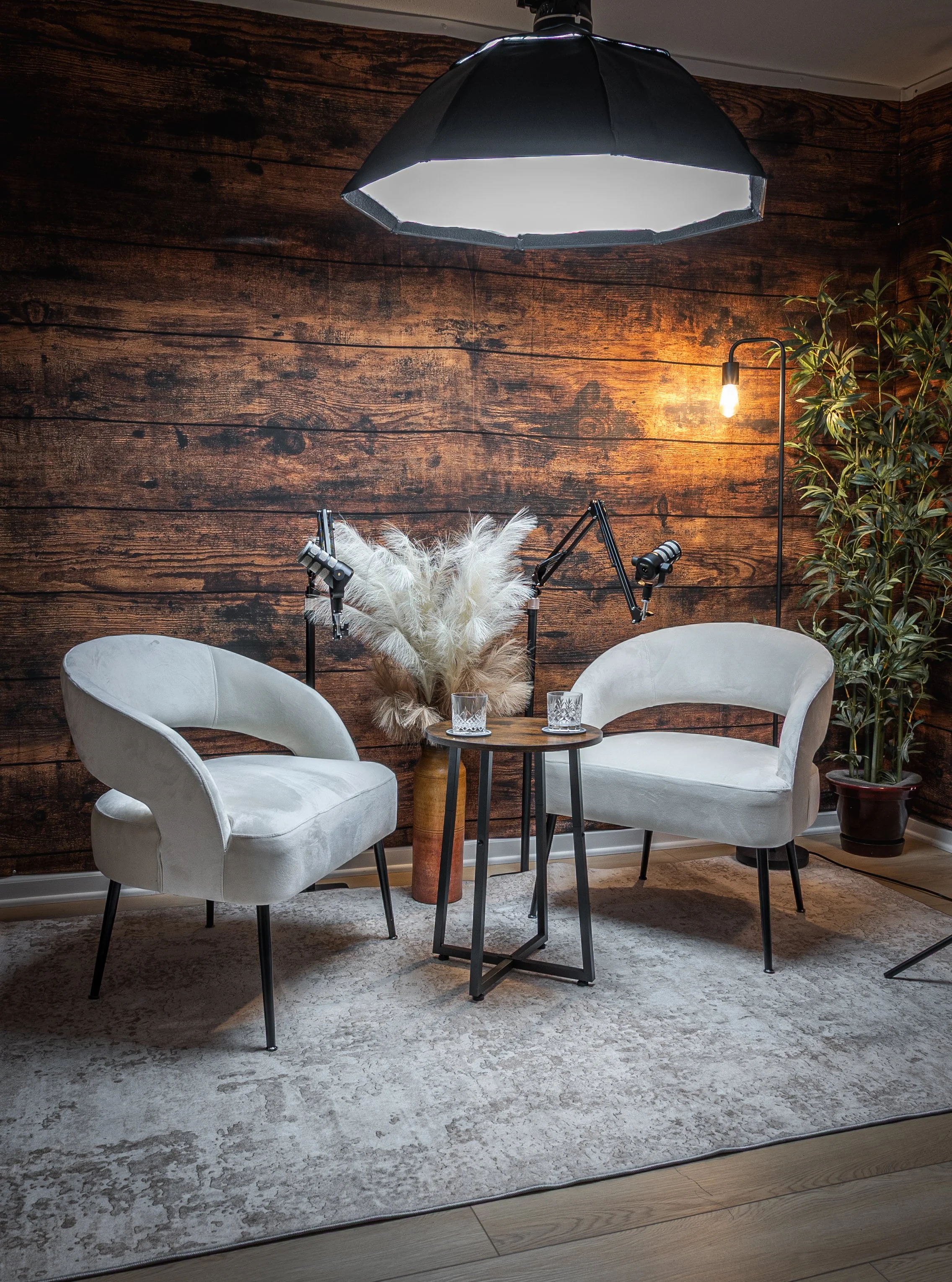 A cozy living room corner featuring two white armchairs, a small round wooden table with two glasses, a floor lamp with an exposed bulb, and potted plant against a wooden plank wall, with professional lighting equipment overhead.