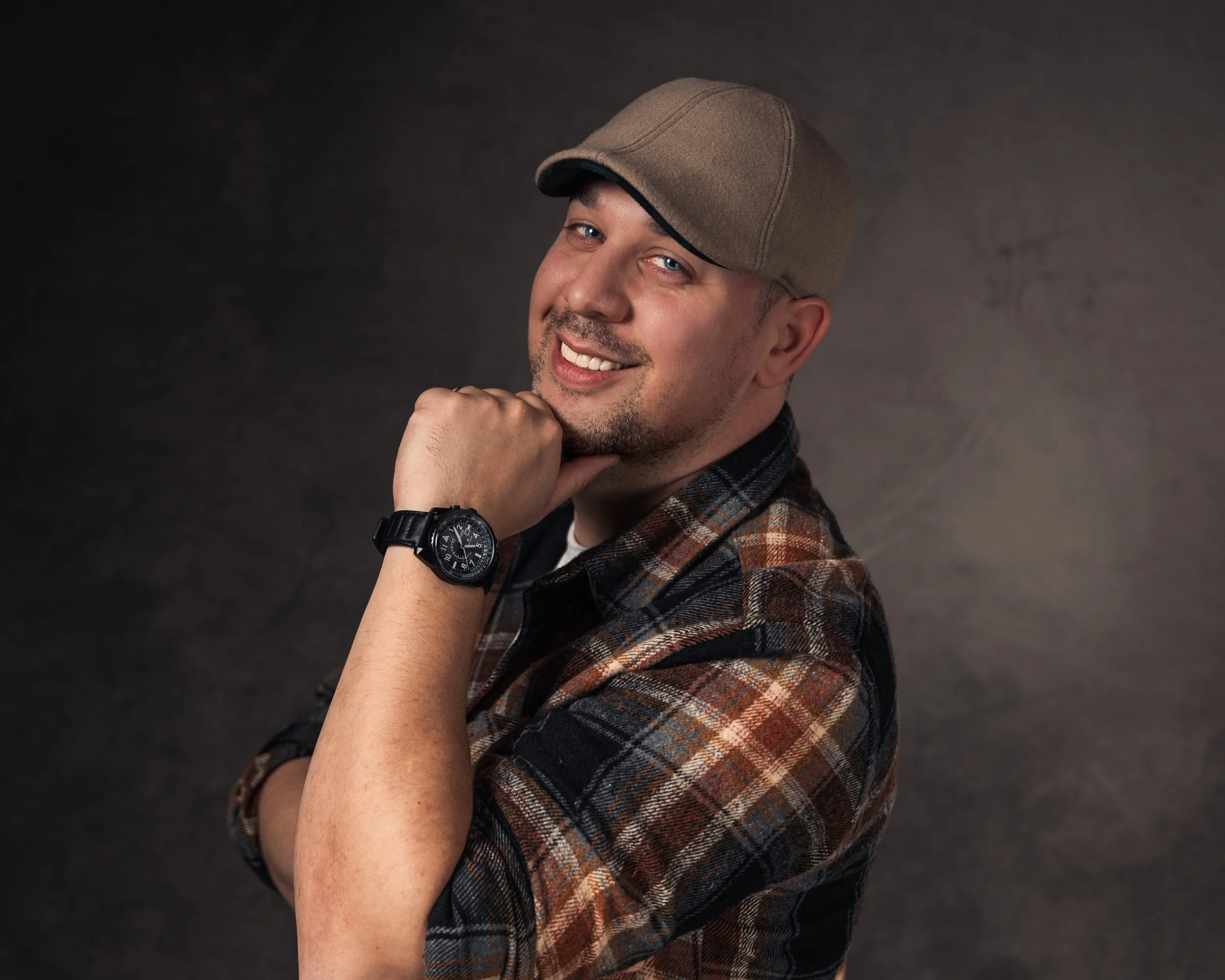 A man smiling with a slight beard, wearing a brown cap, a plaid shirt, and a wristwatch, resting his chin on his hand, against a dark background.