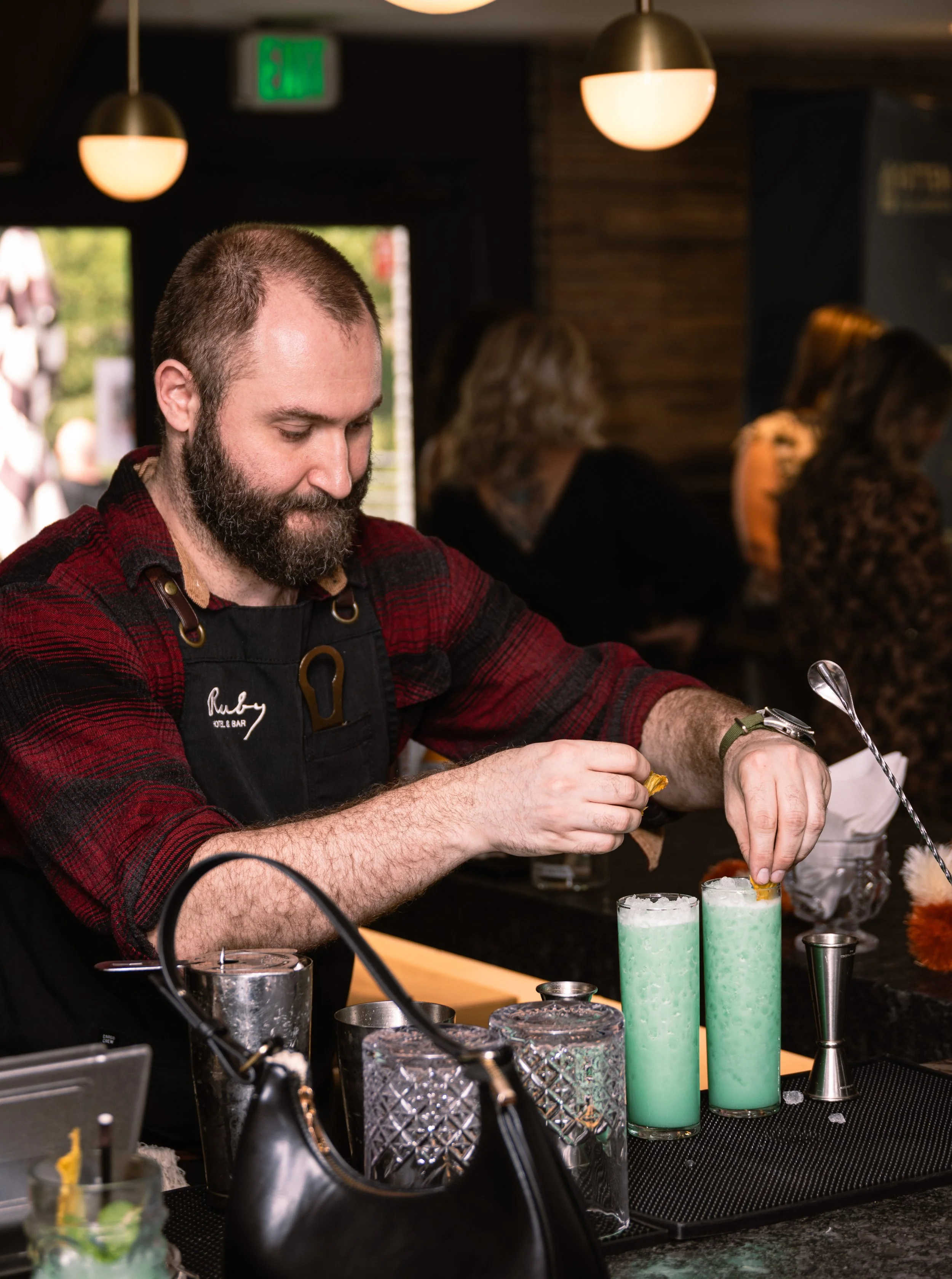 A bartender with a beard and short hair preparing colorful layered drinks at a bar counter, with other patrons in the background.