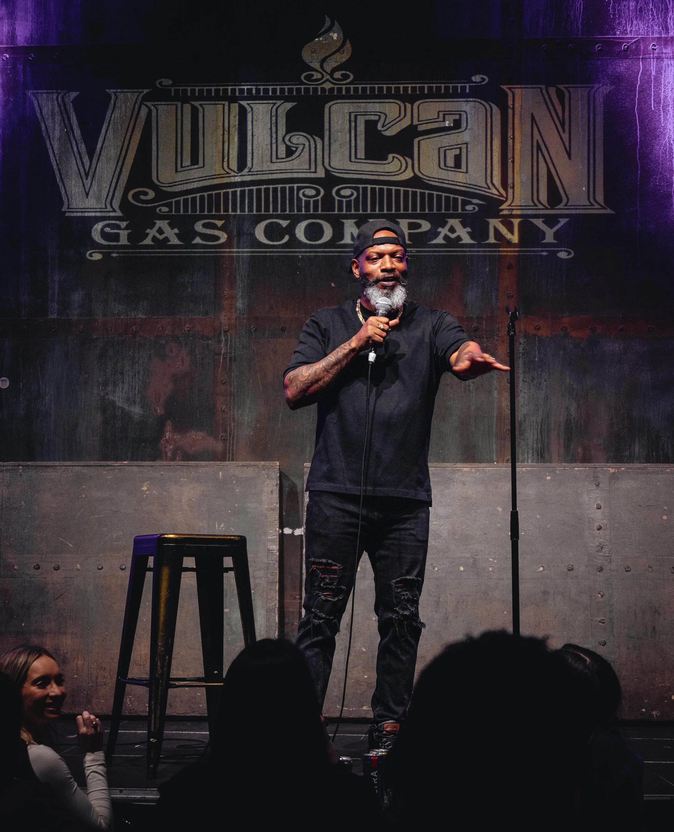 A man with a beard and a backward cap is speaking into a microphone on stage, with a large "Vulcan Gas Company" sign behind him. Audience members in the foreground are watching and smiling.