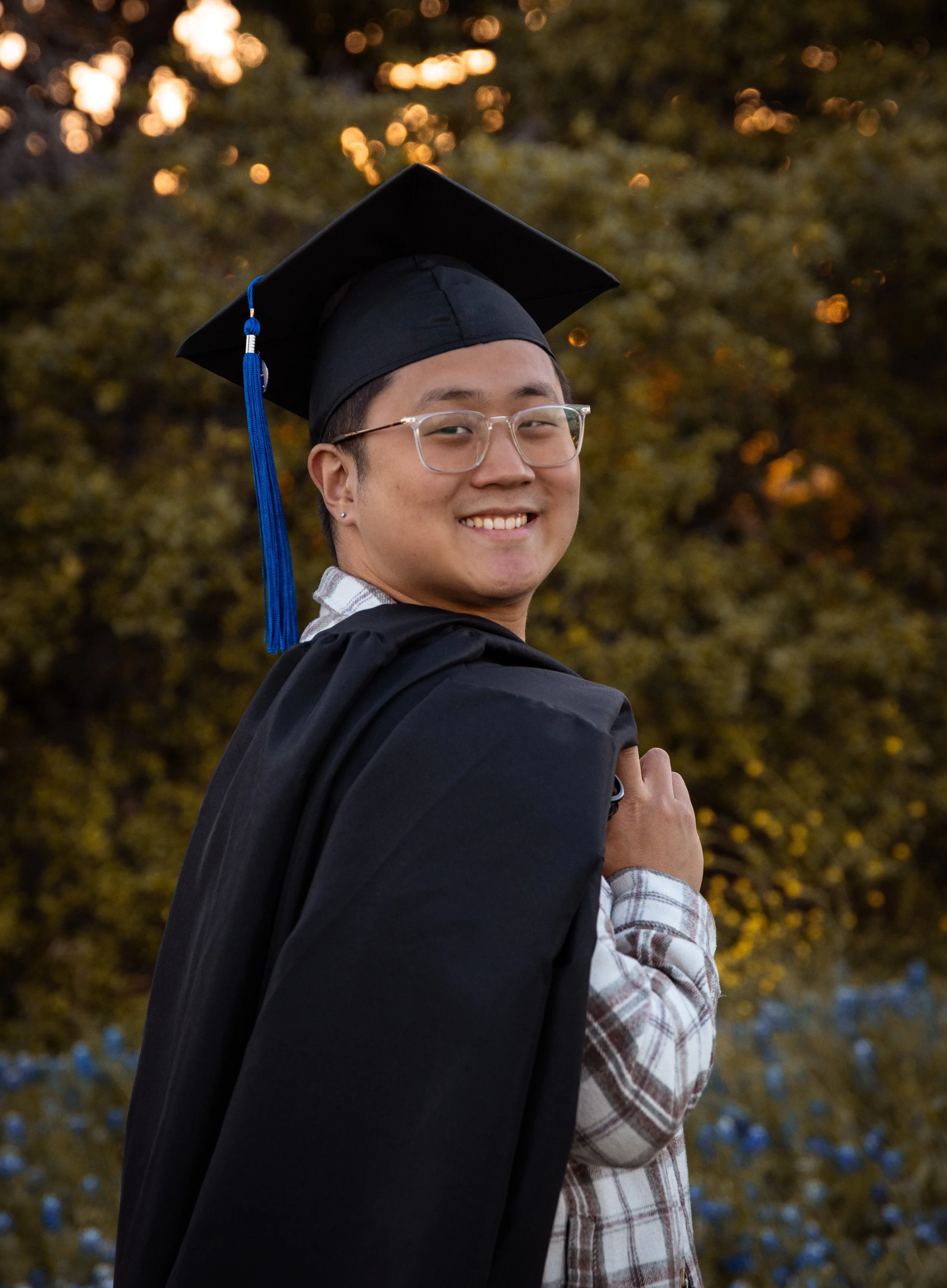 A person smiling at a graduation ceremony, wearing a graduation cap with a blue tassel and gown, outdoors with trees and blurry lights in the background.