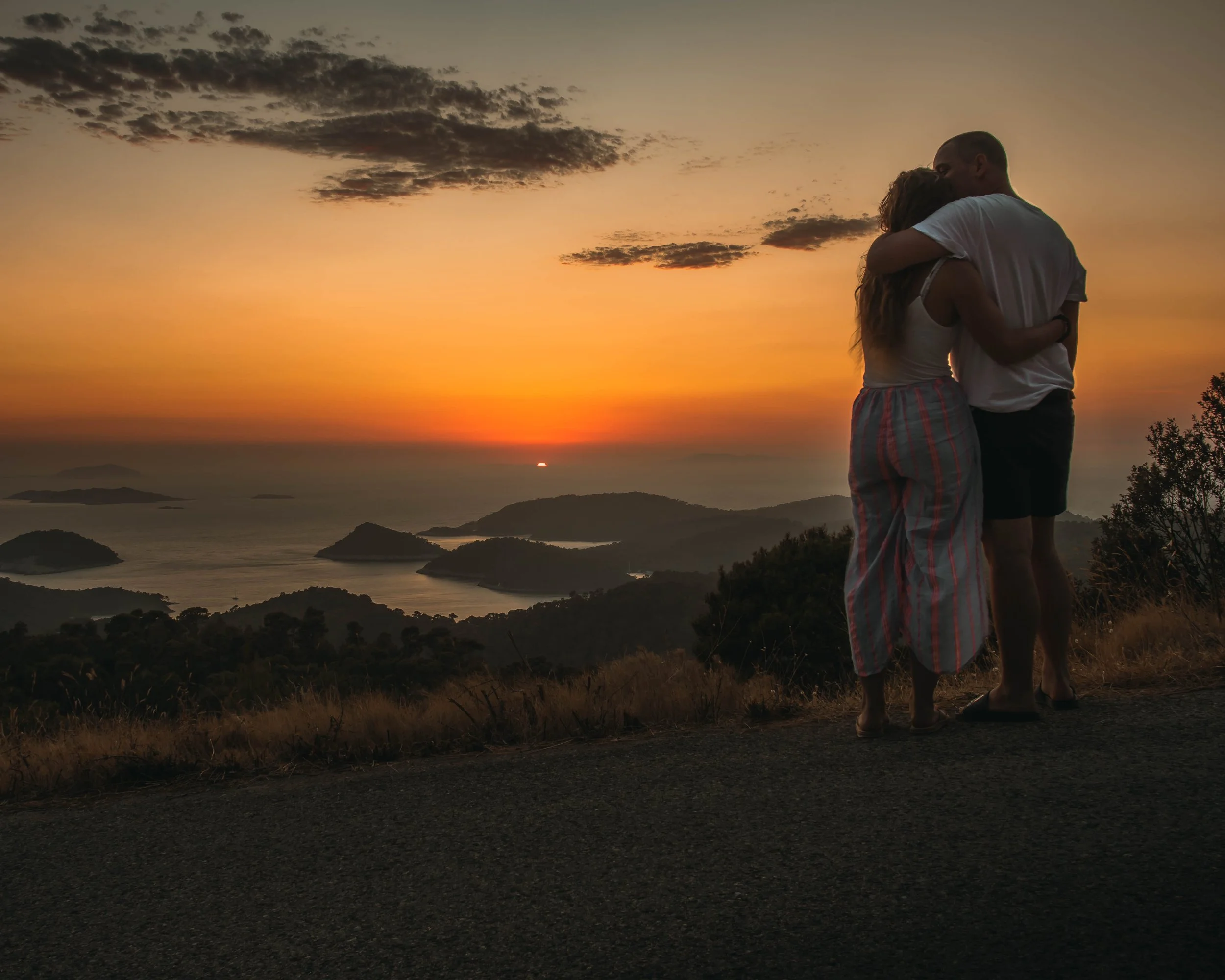 A couple embraces each other at sunset on a hilltop overlooking a body of water with small islands, under a sky with clouds.