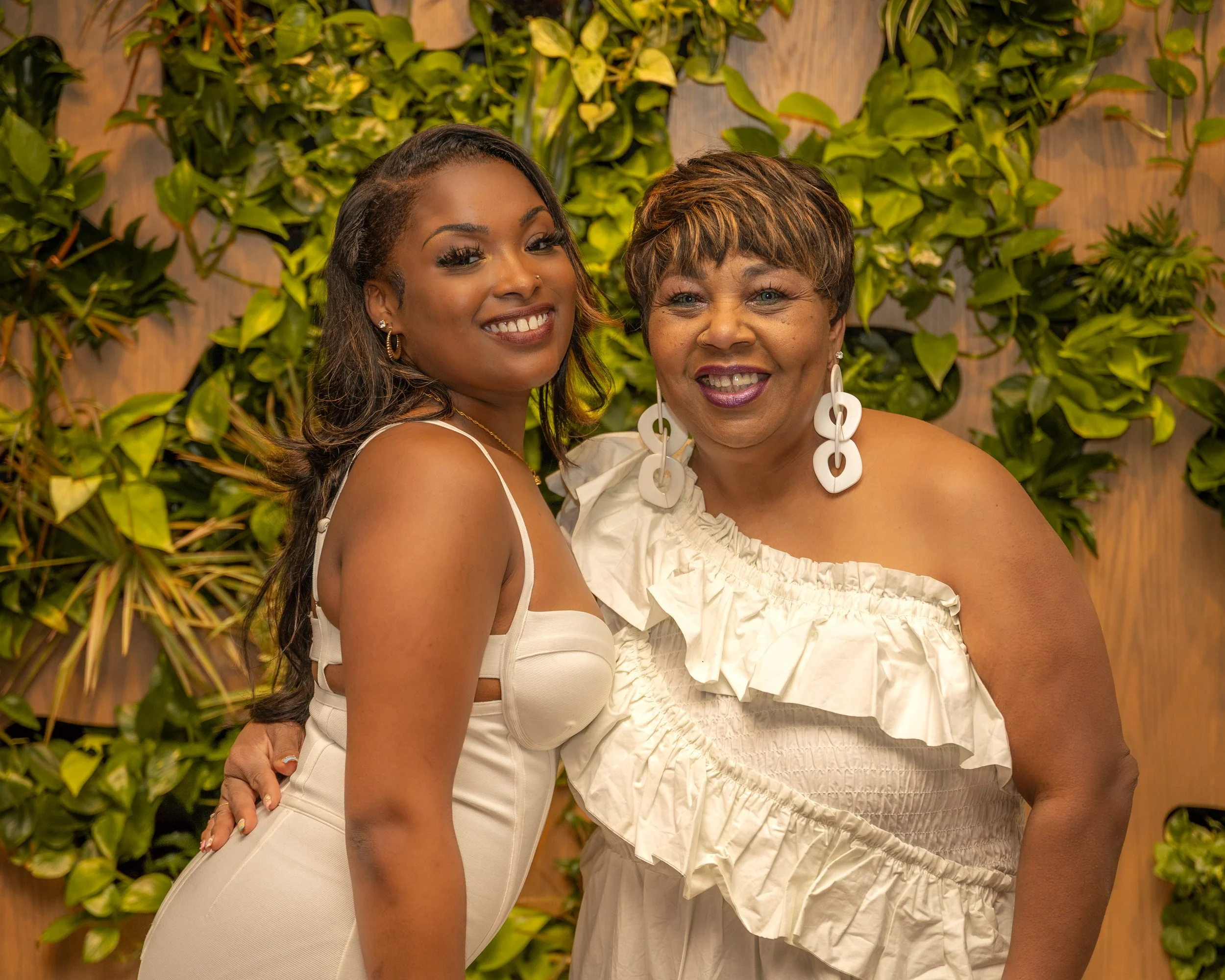 Two women smiling together in front of a green leafy background. The woman on the left has long dark hair, earrings, and is wearing a white dress. The woman on the right has short hair, big earrings, and is wearing a white ruffled top.