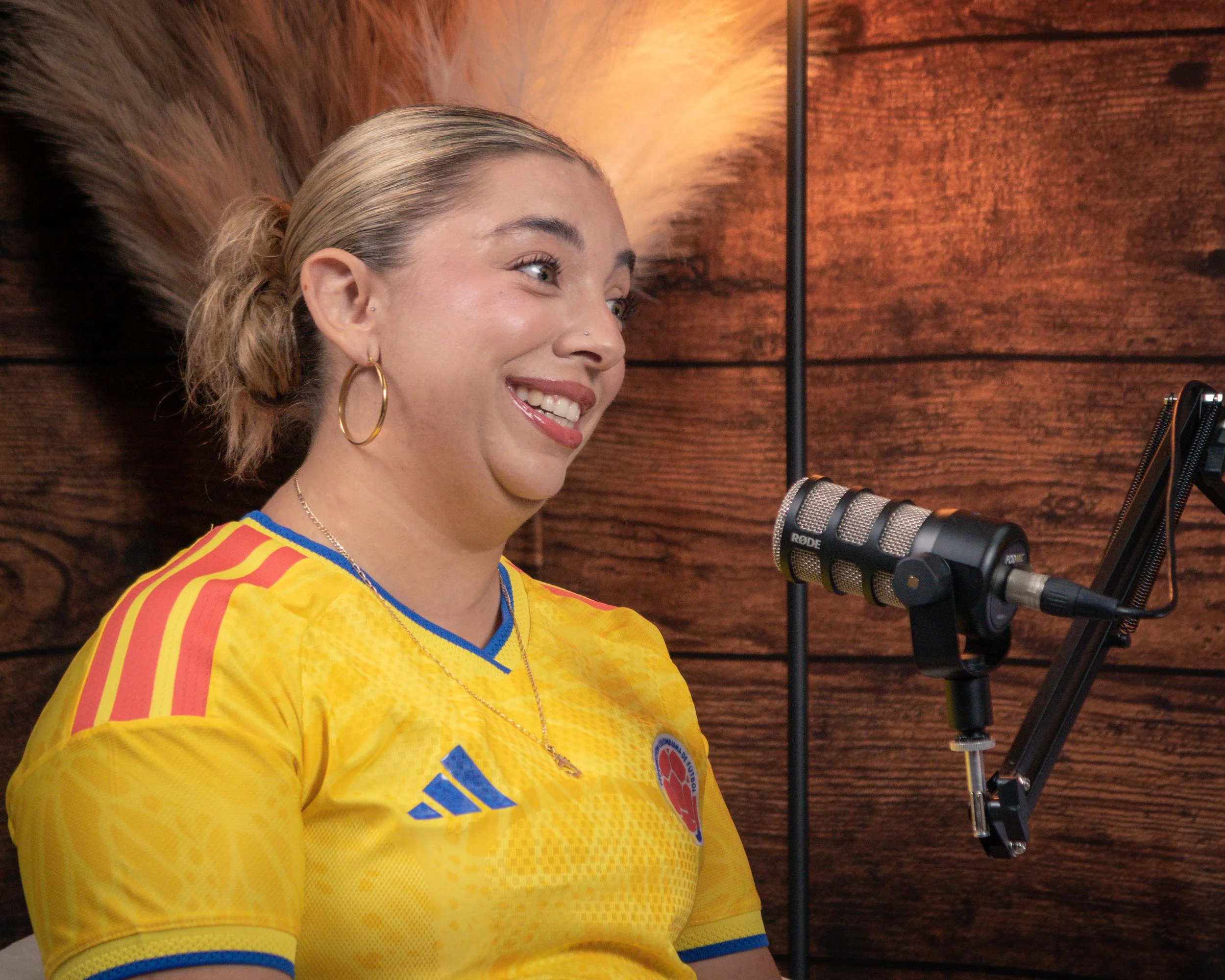 A young woman with blonde hair tied back, wearing a yellow Colombian national soccer team jersey with red, yellow, and blue stripes on the sleeves, smiling and speaking into a professional microphone in a wooden-paneled studio.
