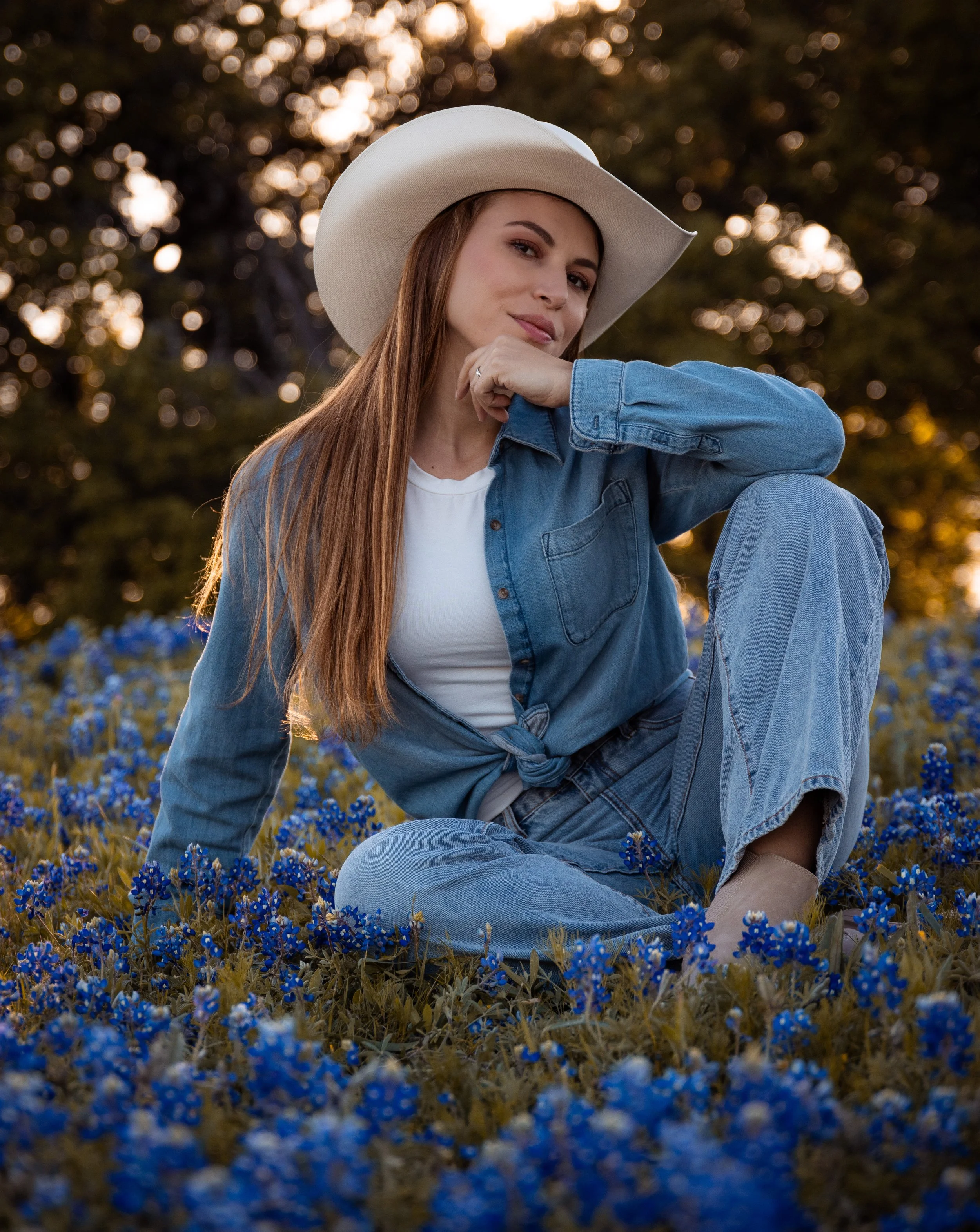 Young woman with long hair wearing a white hat and blue denim jacket sitting in a field of blue flowers during sunset.