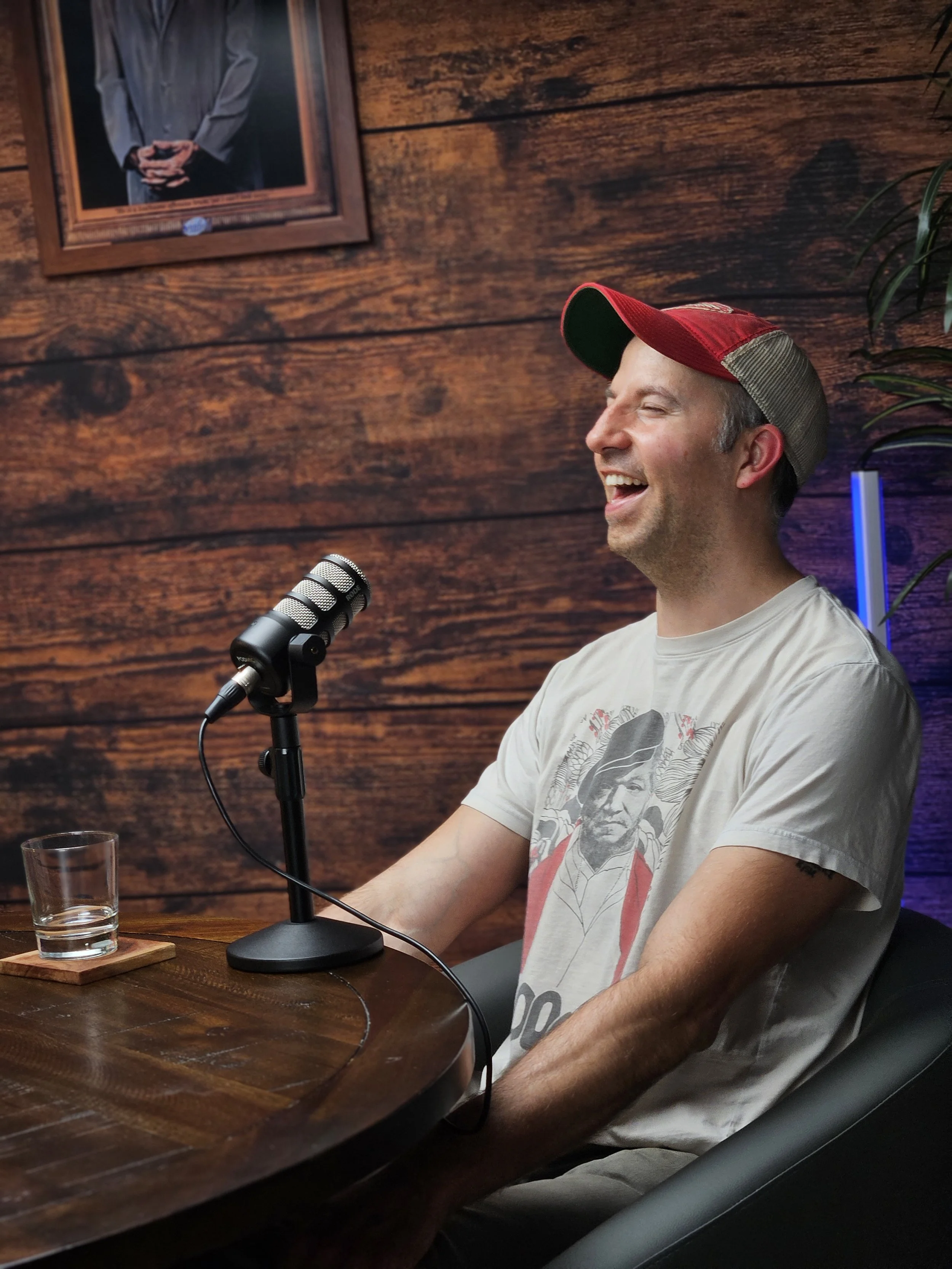 A man wearing a red and gray cap, a white T-shirt with a graphic print, is smiling and sitting at a wooden table with a microphone in front of him. There is a glass of water on the table and a framed portrait of a man on a wooden wall in the backgrou