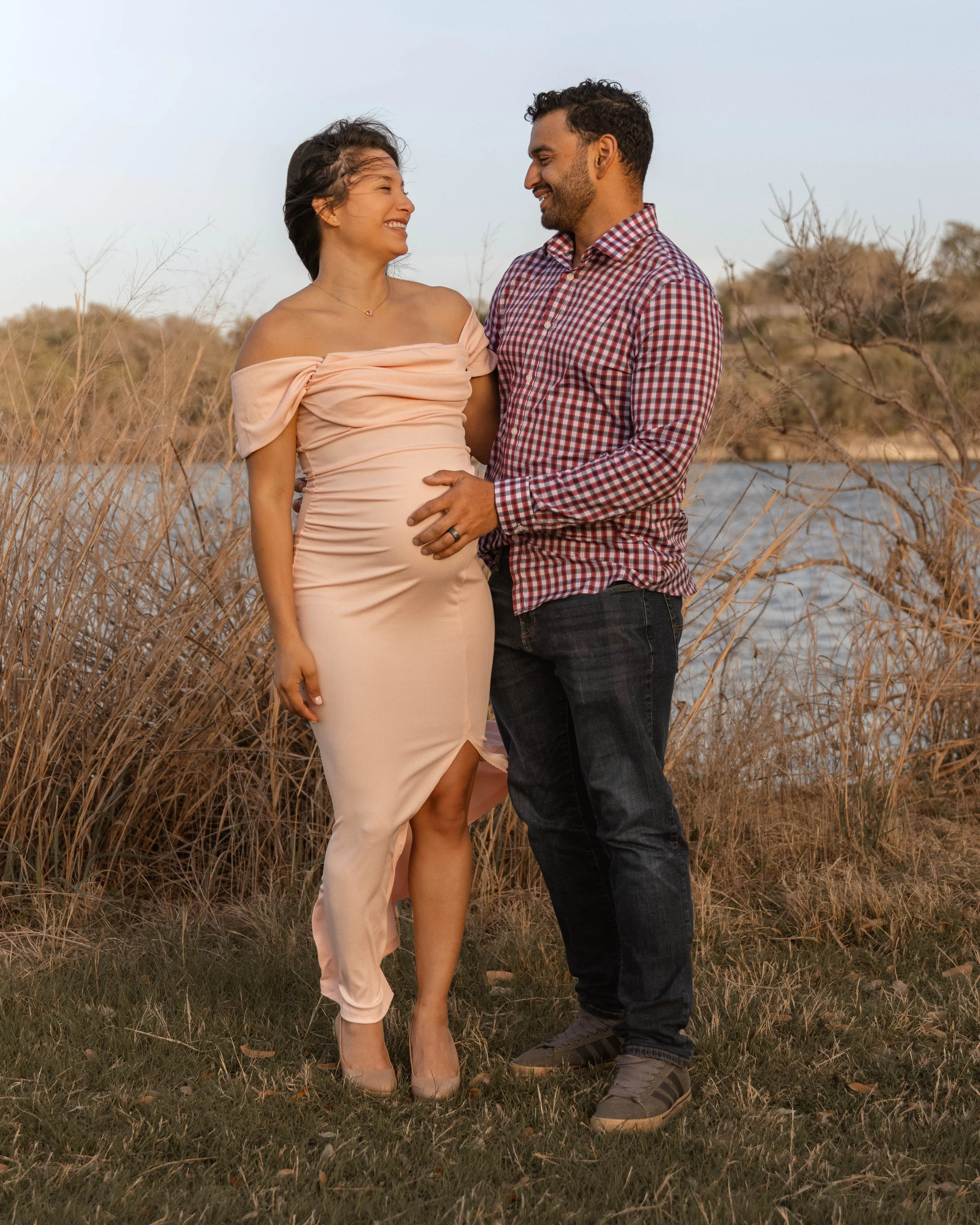 A pregnant woman and a man smiling at each other outdoors near a body of water, surrounded by dry grass and trees, during sunset.