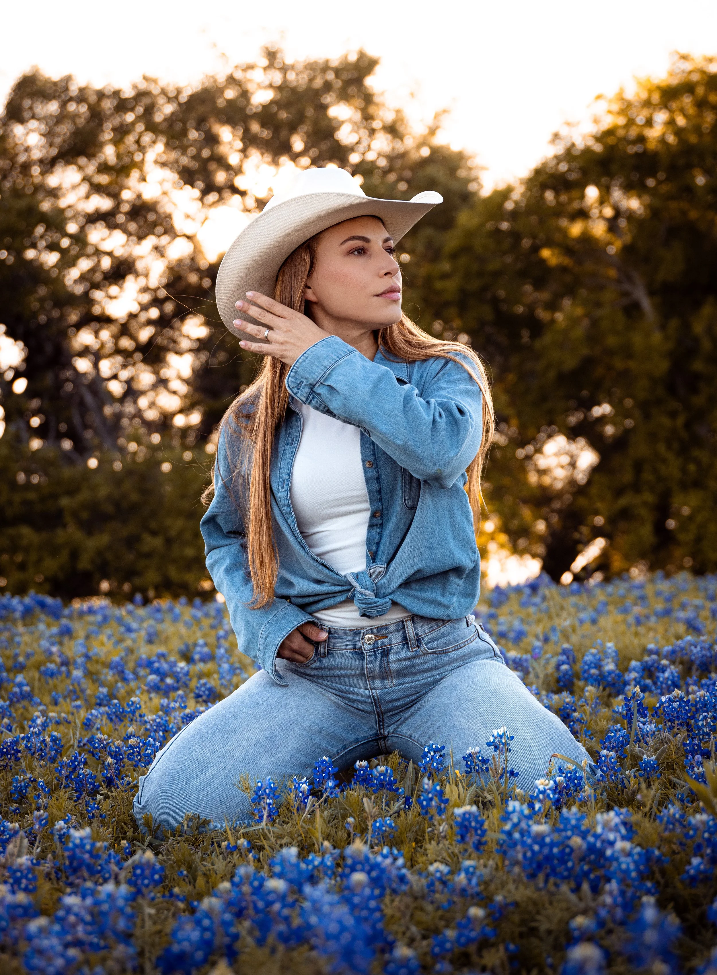 A woman wearing a cowboy hat, denim jacket, and white shirt kneels in a field of blue flowers, with the sunset and trees in the background.
