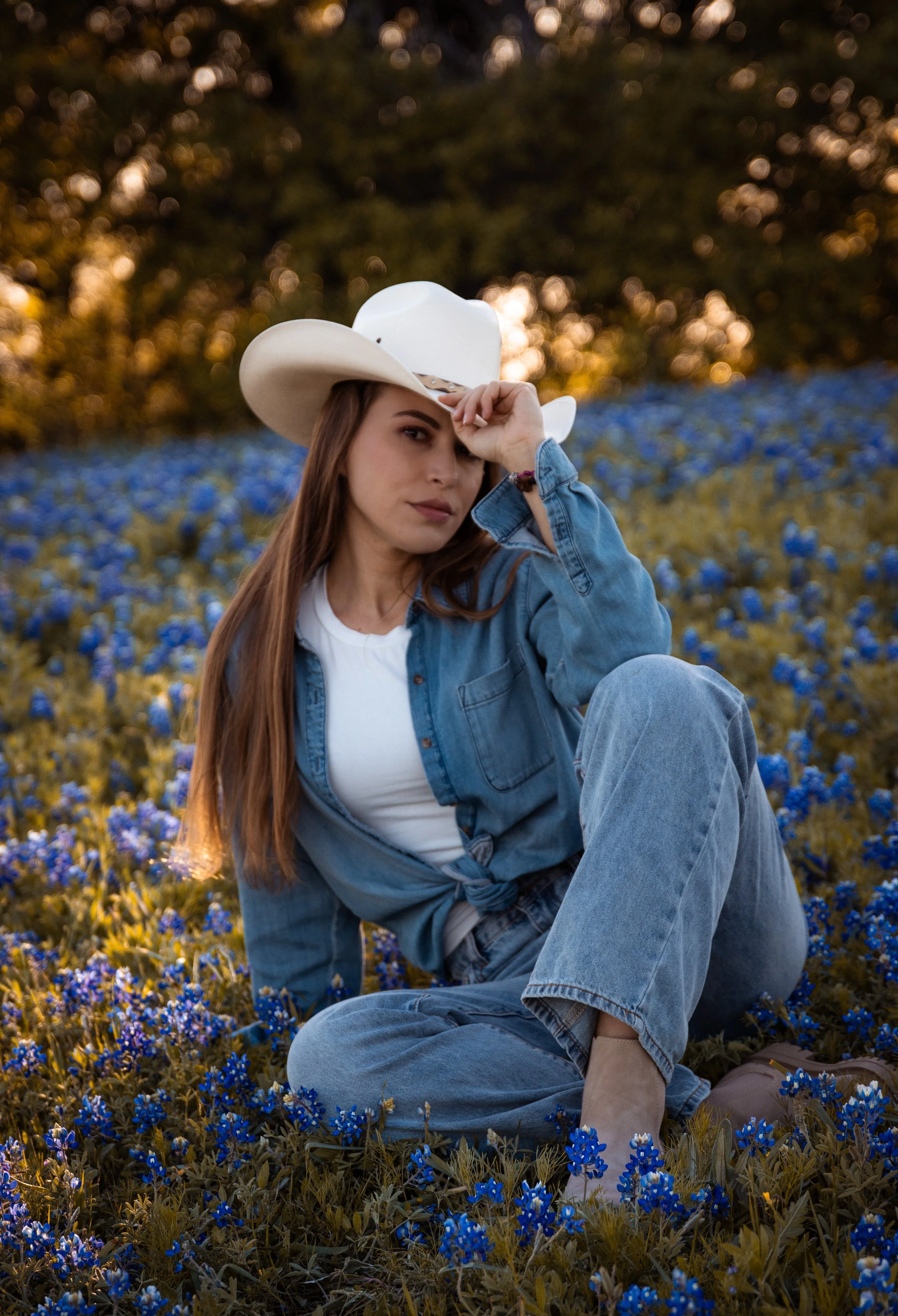 A woman with long brown hair and fair skin sitting in a field of blue flowers at sunset. She is wearing a white cowboy hat, a white shirt, a denim jacket, and jeans. She is looking at the camera and holding the brim of her hat with one hand. The back