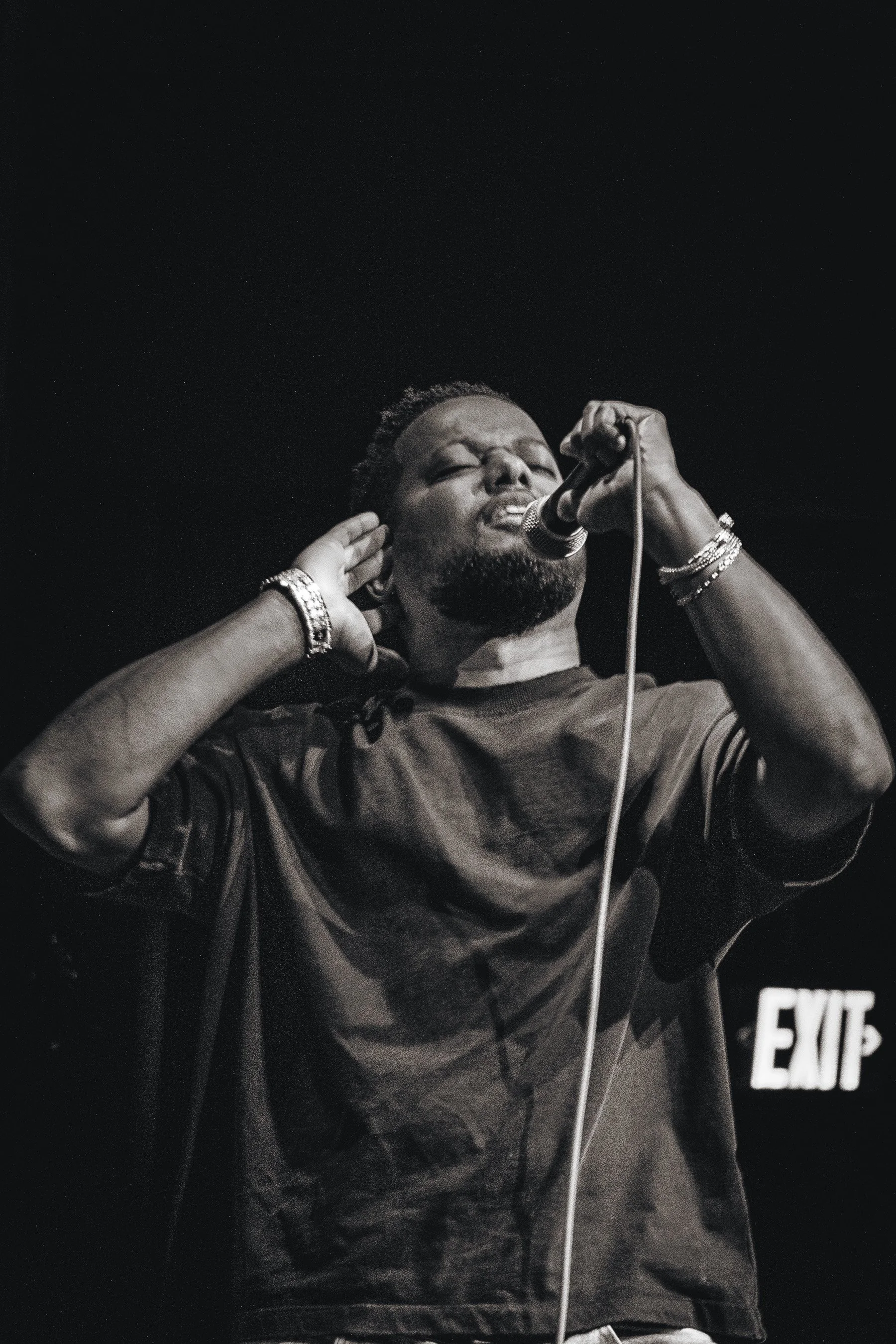 A person singing into a microphone with hands near ears, wearing bracelets, and standing in front of an exit sign.