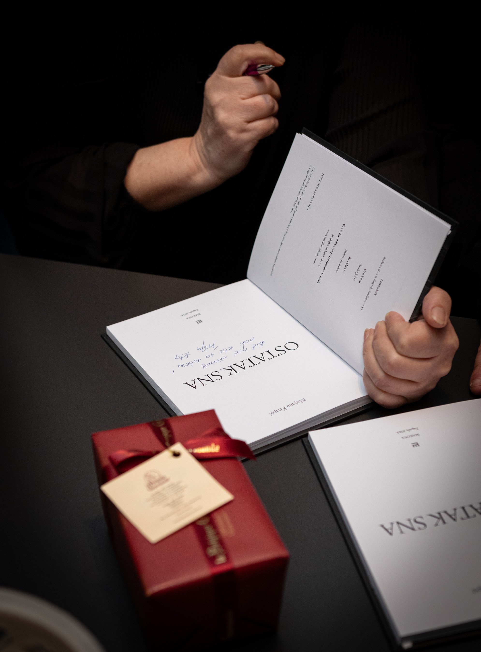 A person holding an opened book or program with the title 'Osteansha'. The individual is wearing a dark long sleeve shirt, and the background is black. There's a red wrapped gift and a small white card with a gold label on the table in front of them.