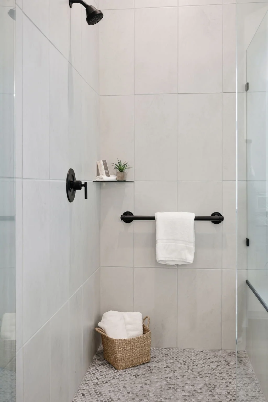 A modern walk-in shower with white tiled walls, black fixtures, a glass door, a towel bar with a white towel, a small shelf with a plant and phone, and a basket with a white towel on the pebblestone floor.