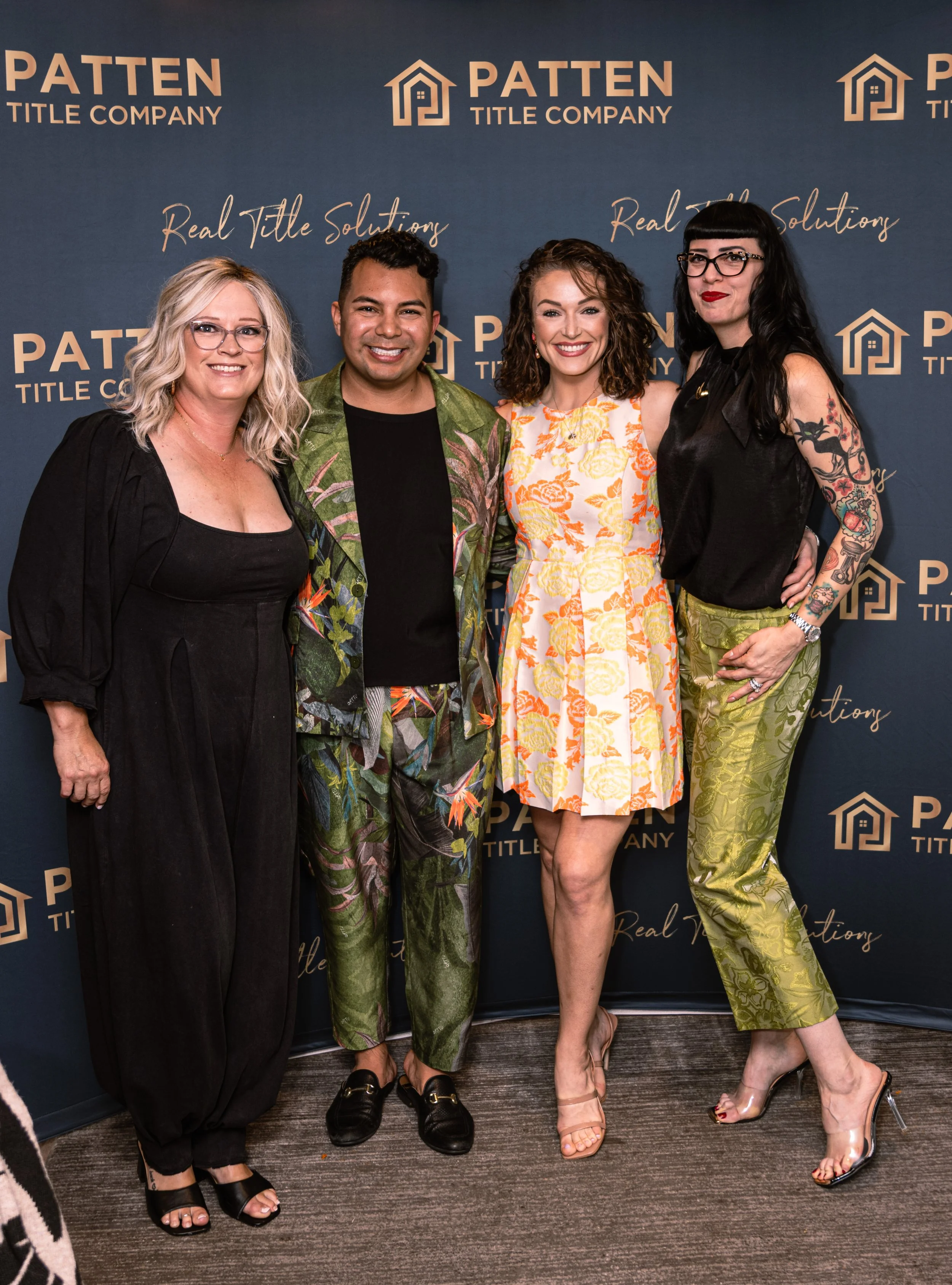 Four people standing side by side smiling in front of a blue backdrop with logos and phrases, dressed in colorful clothing, at a professional event.