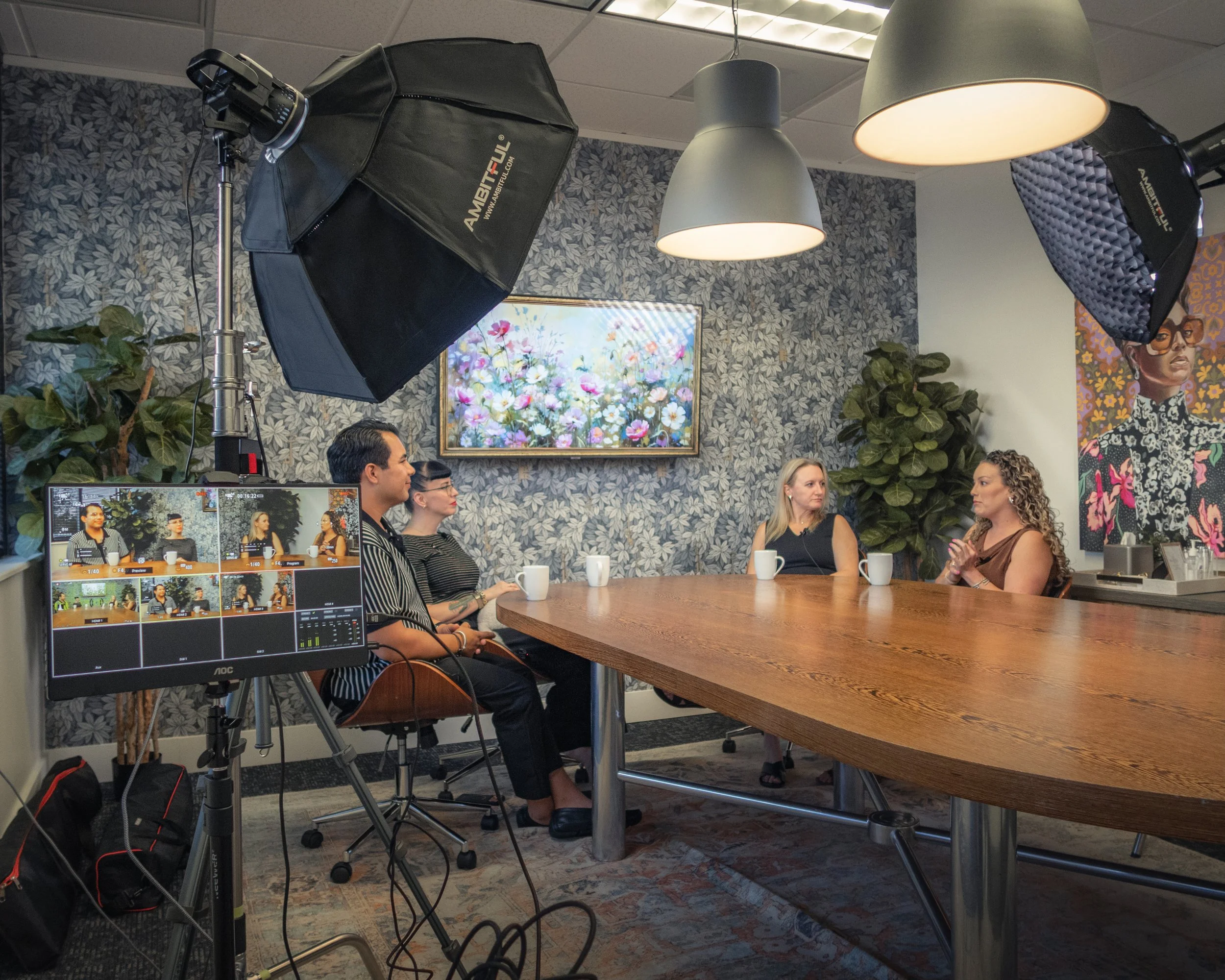 A group of four people sitting around a wooden conference table in a room with floral patterned wallpaper, listening to a woman speaking. There are cameras, lighting equipment, and a monitor showing three of the people.