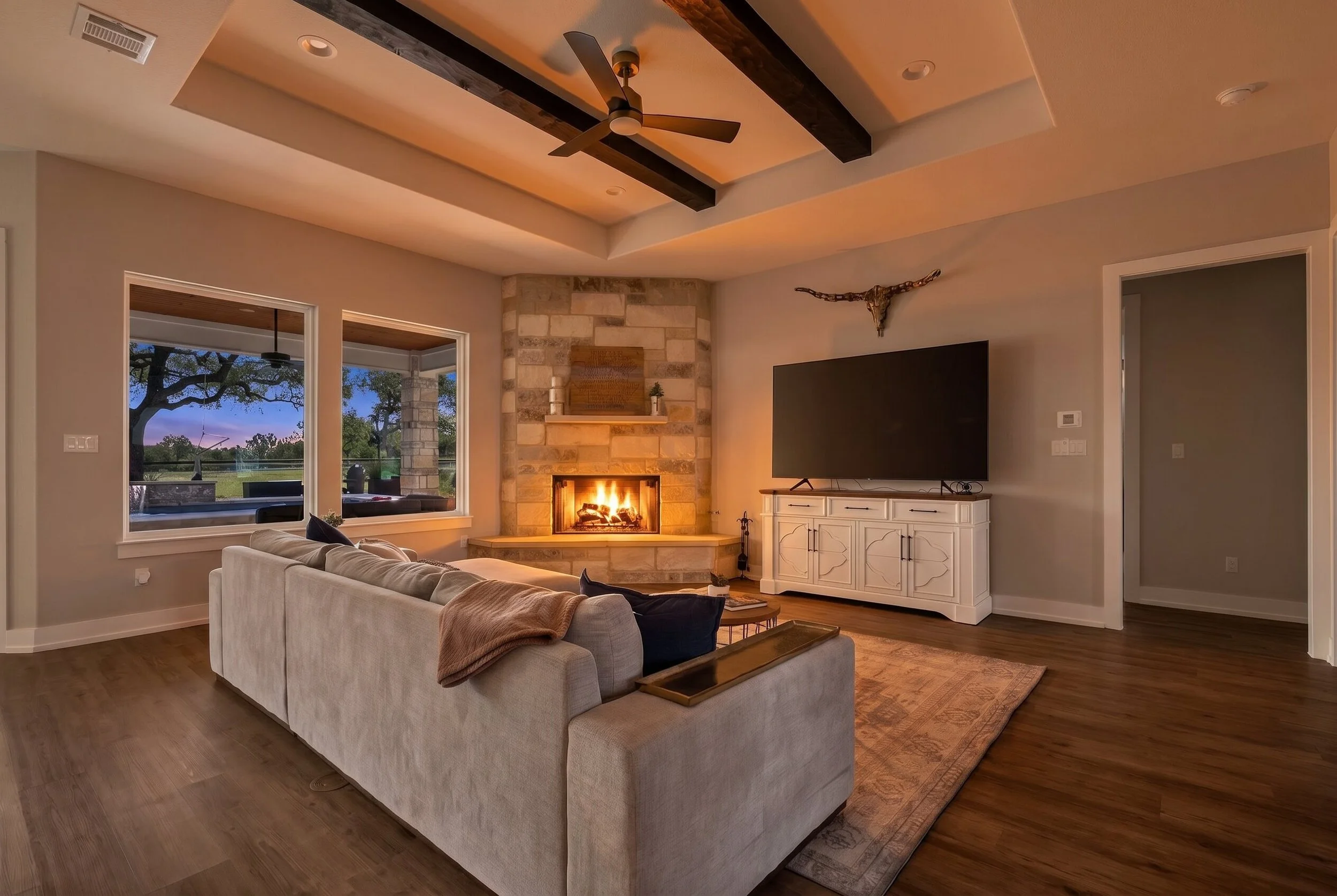 Living room with a beige sofa, a stone fireplace with a fire burning, a white TV cabinet with a large flat-screen TV, a mounted animal skull with horns on the wall, large windows showing outdoor trees and a sunset sky, ceiling fan, hardwood floors, cozy and warm ambiance.