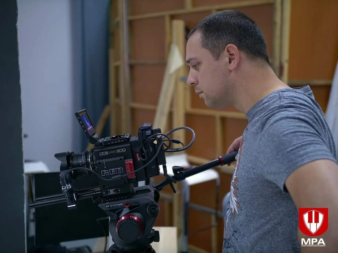 A man in a grey T-shirt operating a professional RED digital cinema camera on a tripod, in what appears to be a film or video production set with wooden shelves and equipment in the background.