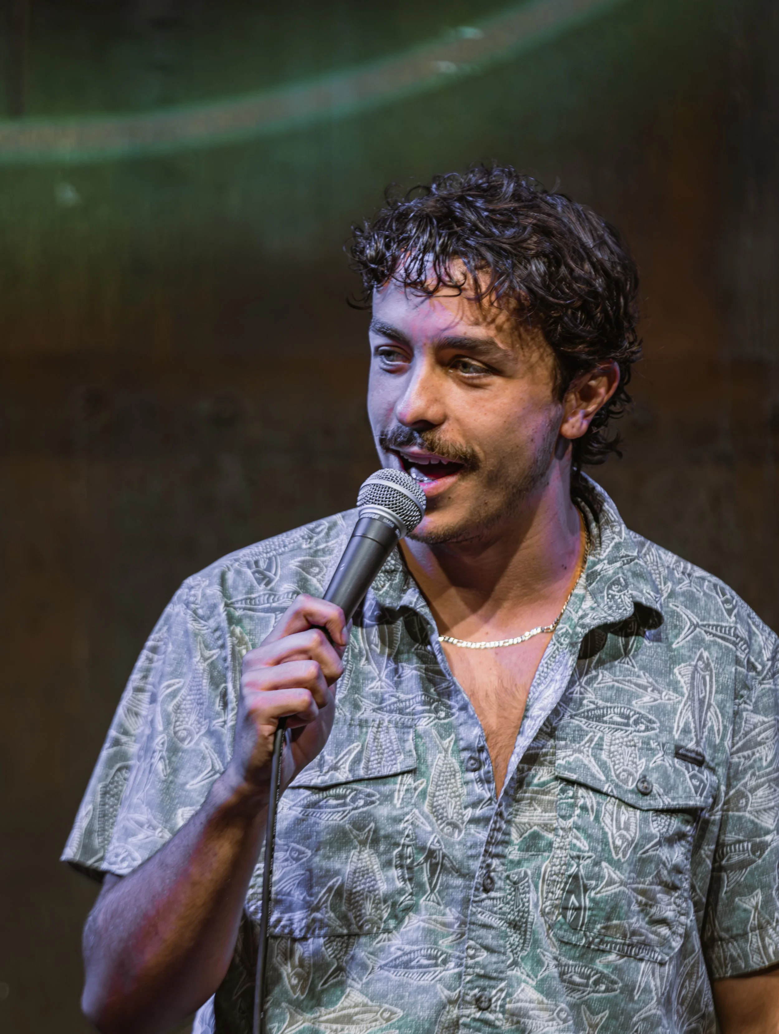 A young man with curly brown hair, a mustache, and a beard is holding a microphone and speaking. He is wearing a patterned shirt with an open collar and a gold chain necklace. The background is dark and blurred.