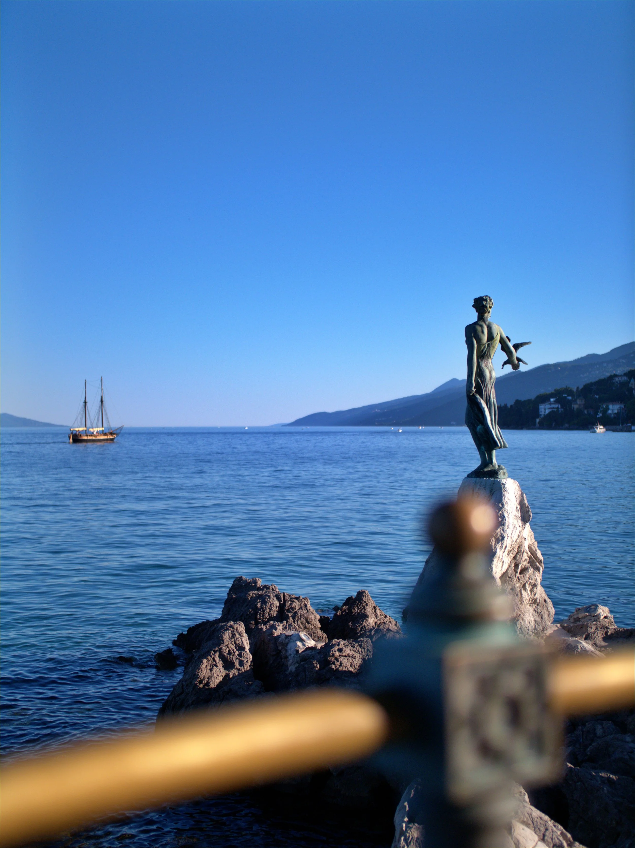 A scenic view of a harbor with a statue of a woman on a rock in the foreground, a sailboat on the water, and mountains in the distance under a clear blue sky.